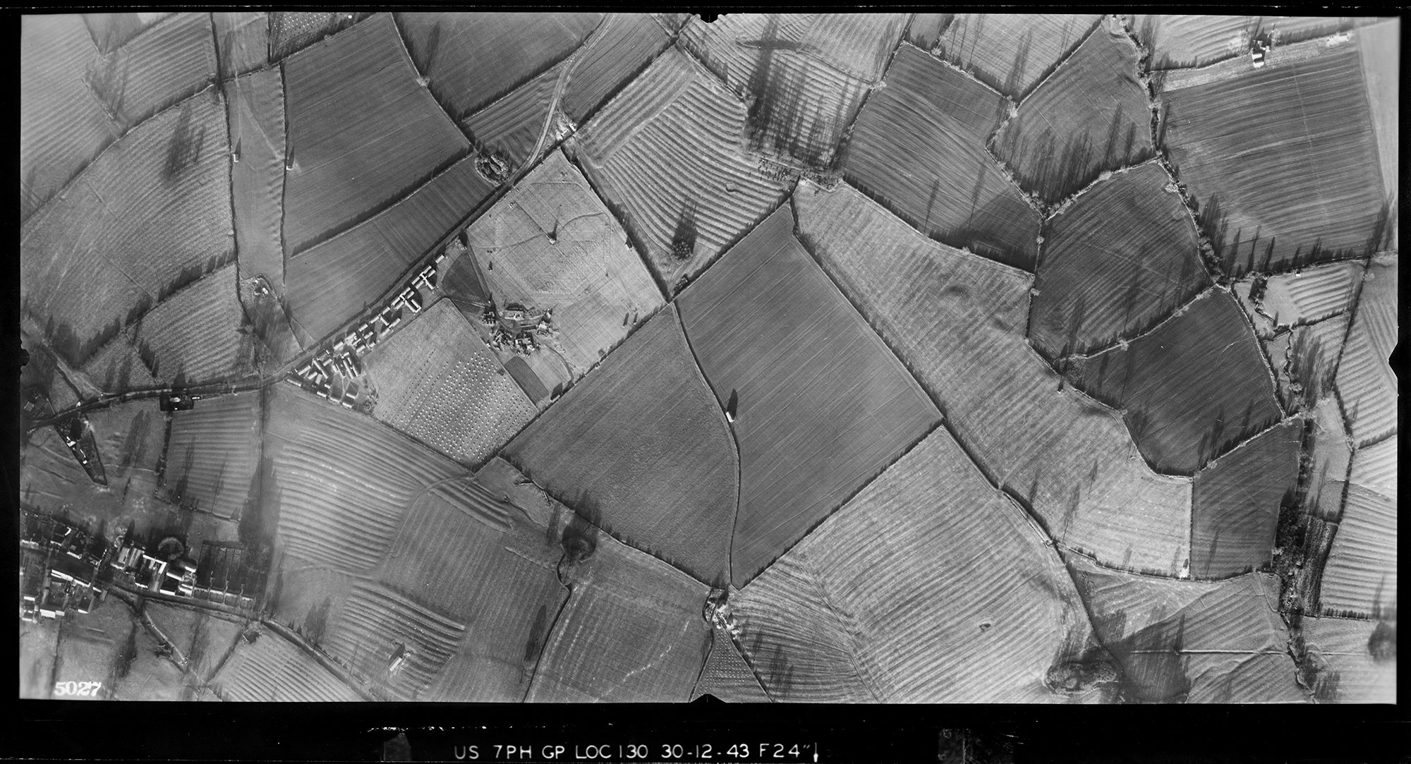 A black and white vertical aerial photograph showing a patchwork of irregular fields bounded by hedges and tracks. Several of the fields contain parallel lines of ridge and furrow ploughing.