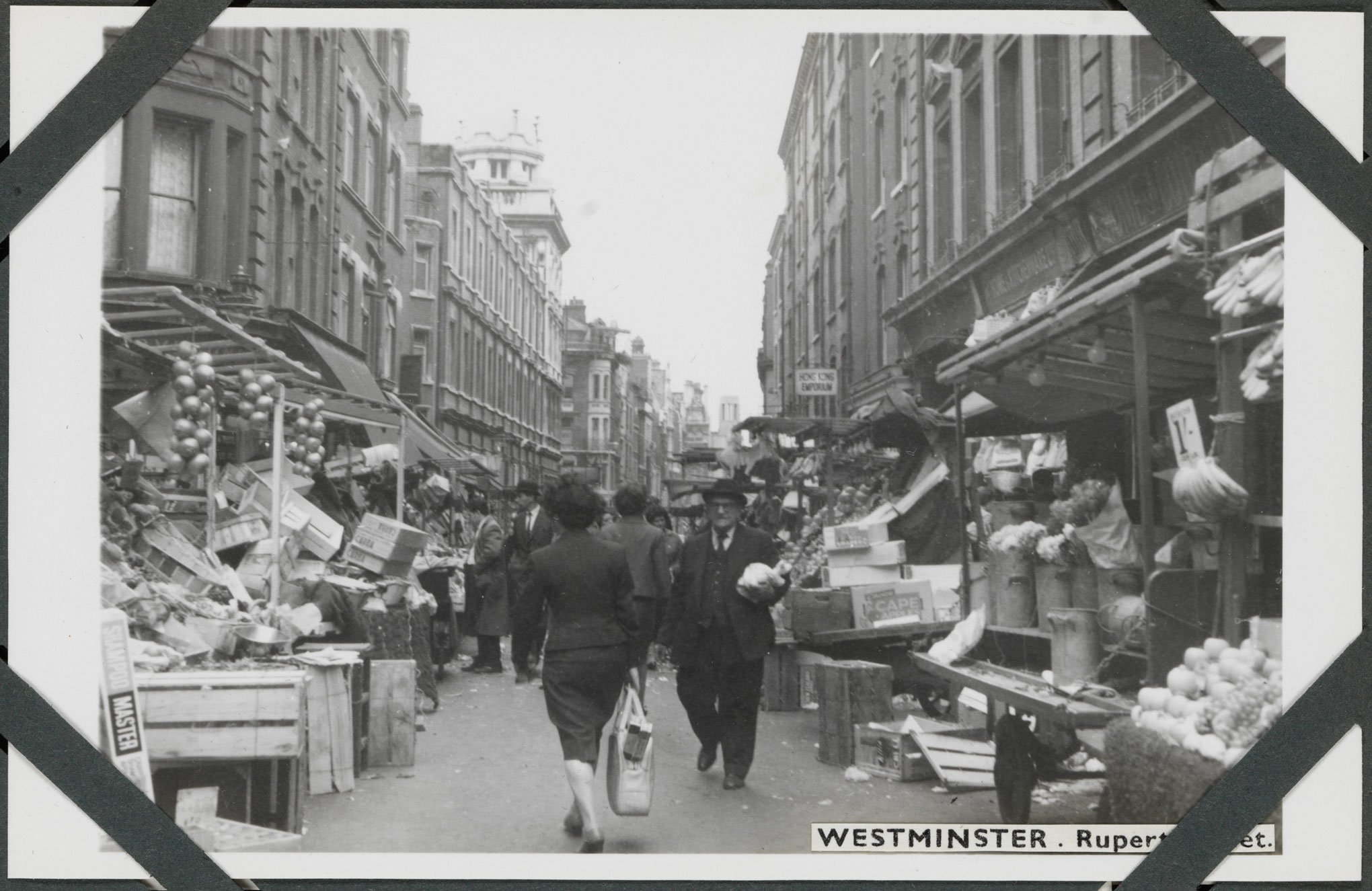 Black and white photograph of an urban street scene featuring people walking in a road flanked by market stalls set out in front of rows of stone-fronted buildings of multiple storeys. 