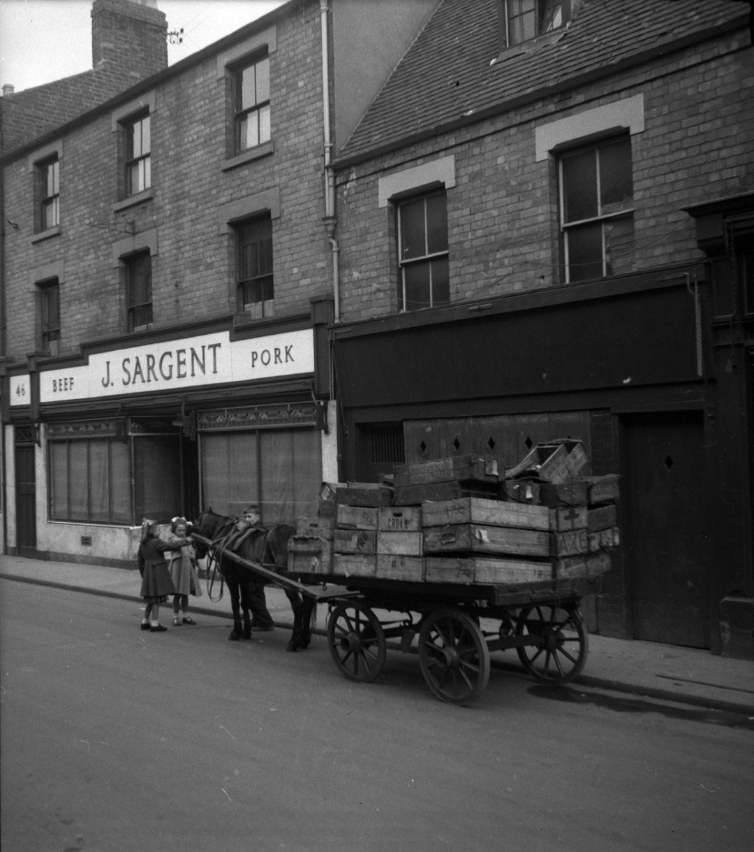 A black and white photograph of a street with brick buildings and old shop fronts with a horse and cart into front of them.
