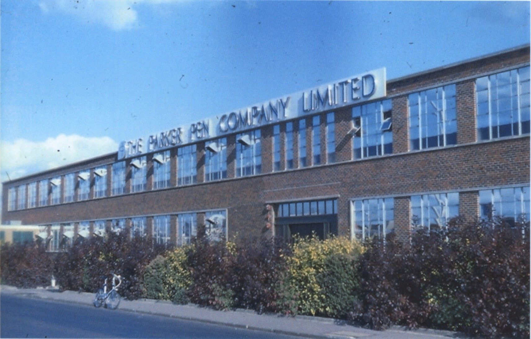 A photograph of a factory building with many glass windows. A sign at the top of the building reads "THE PARKER PEN COMPANY LIMITED".