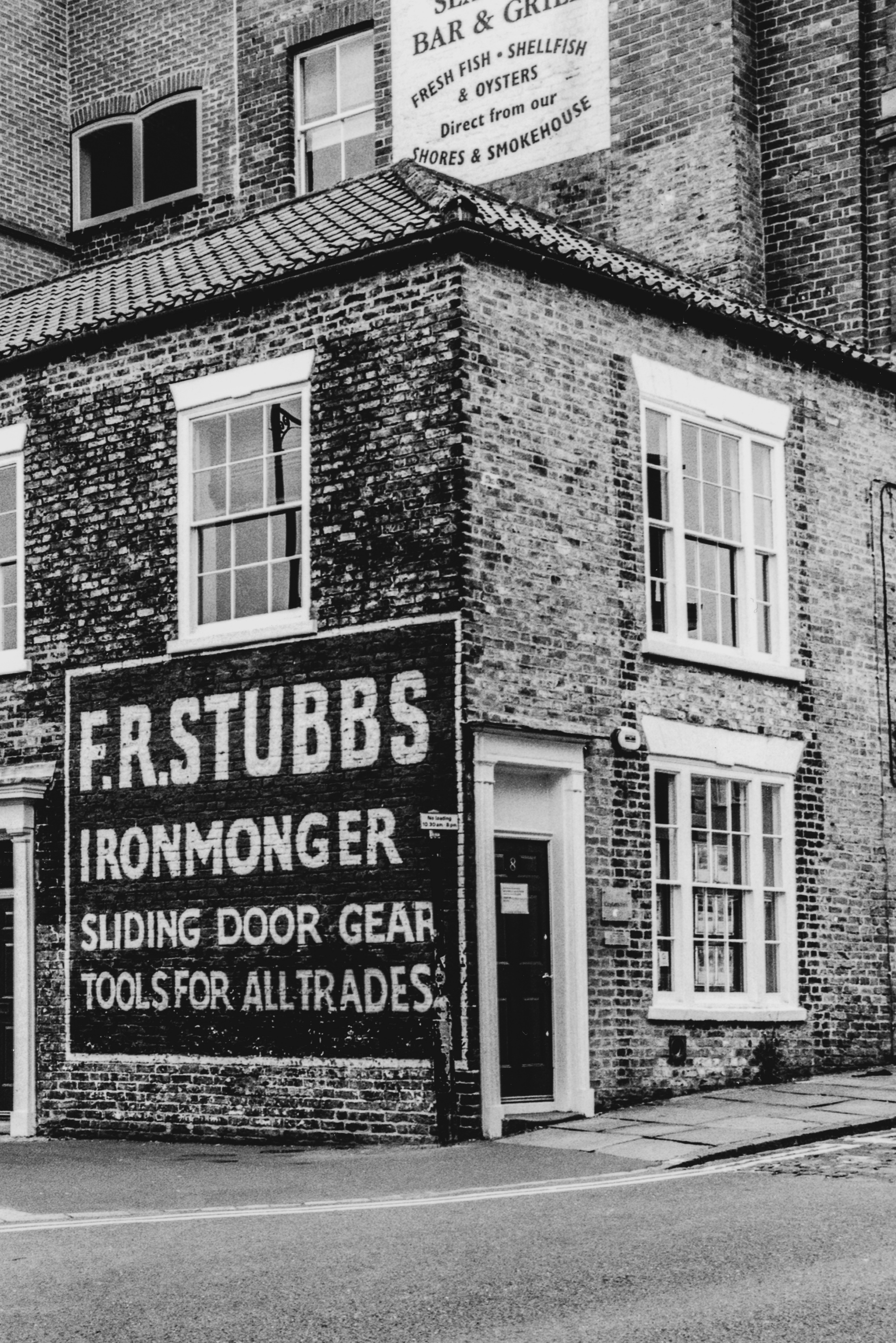 A black and white photograph of the exterior of a brick building. A painted sign with a dark background and white lettering reads: "F.R. STUBBS; IRONMONGER; SLIDING DOOR GEAR; TOOLS FOR ALL TRADES"
