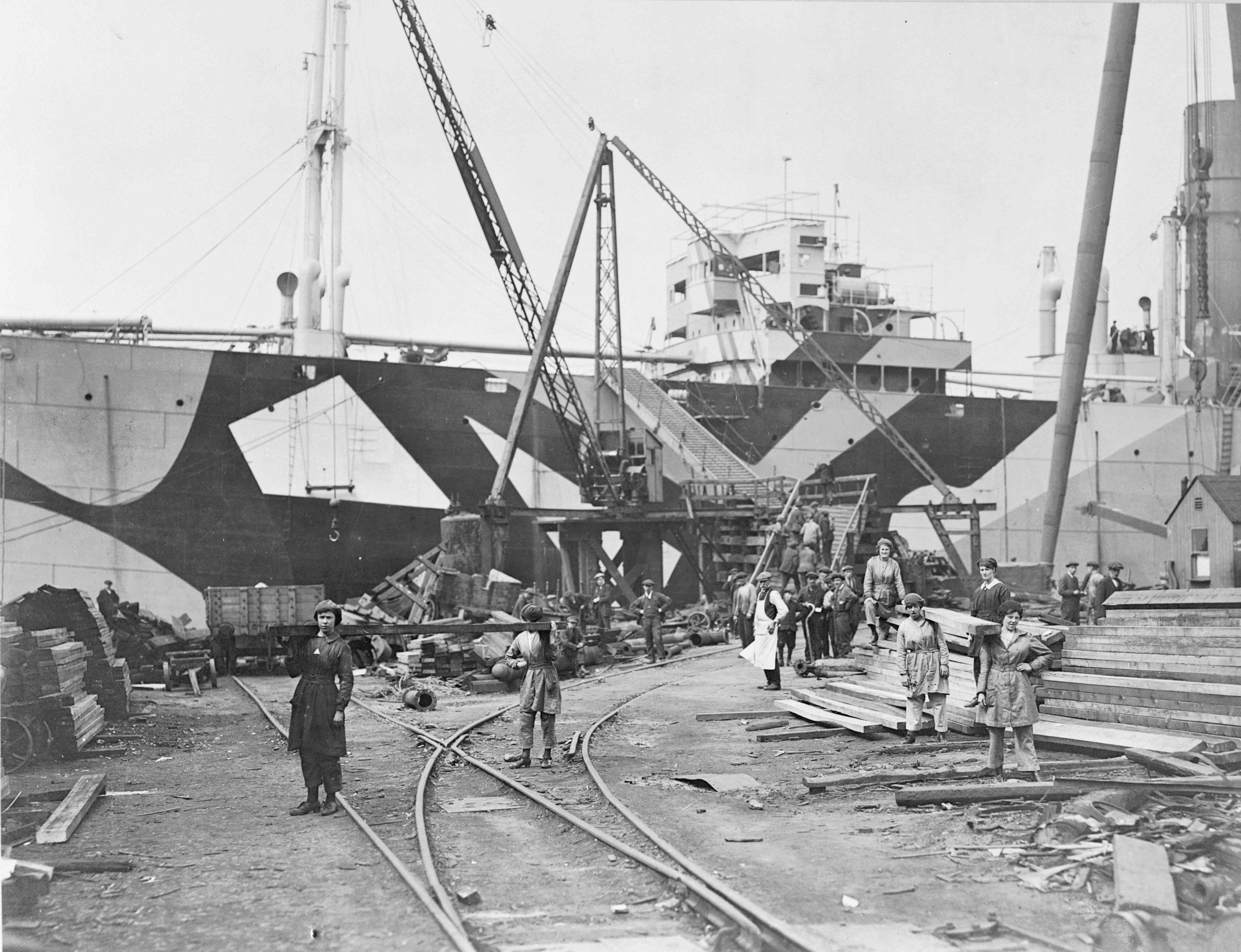 A black and white image of a shipbuilding yard with a boat in the background and women carrying timber. 