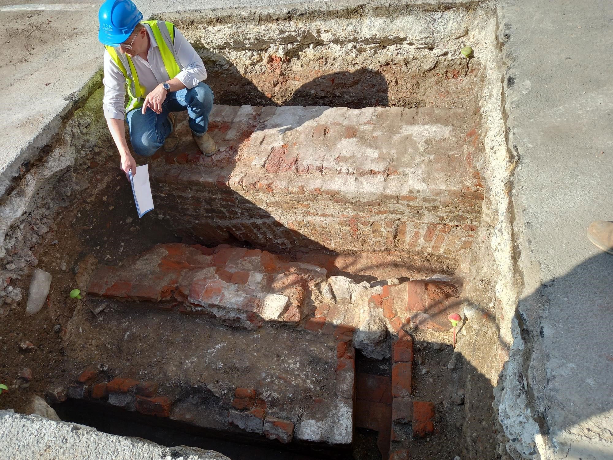 An archaeologist kneeling on a brick arch pointing at earlier masonry within a small archaeological trench.