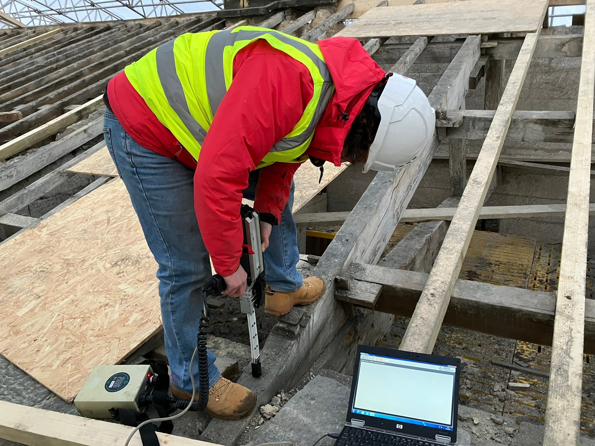 Photograph showing dark-haired woman in red coat, high vis and hard hat using a decay detection drill on Belsay Hall roof trusses. 