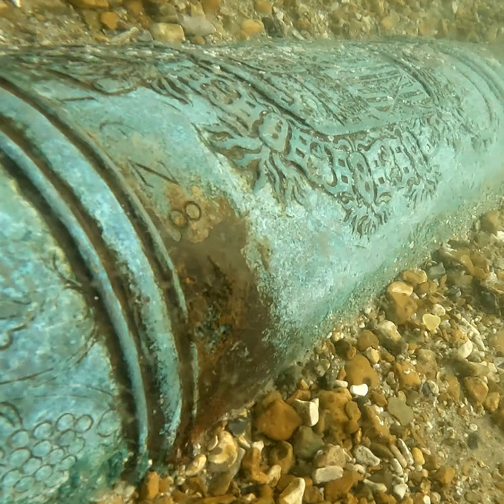 Green metallic cannon inscribed with patterns and text, sitting in a layer of gravel underwater.