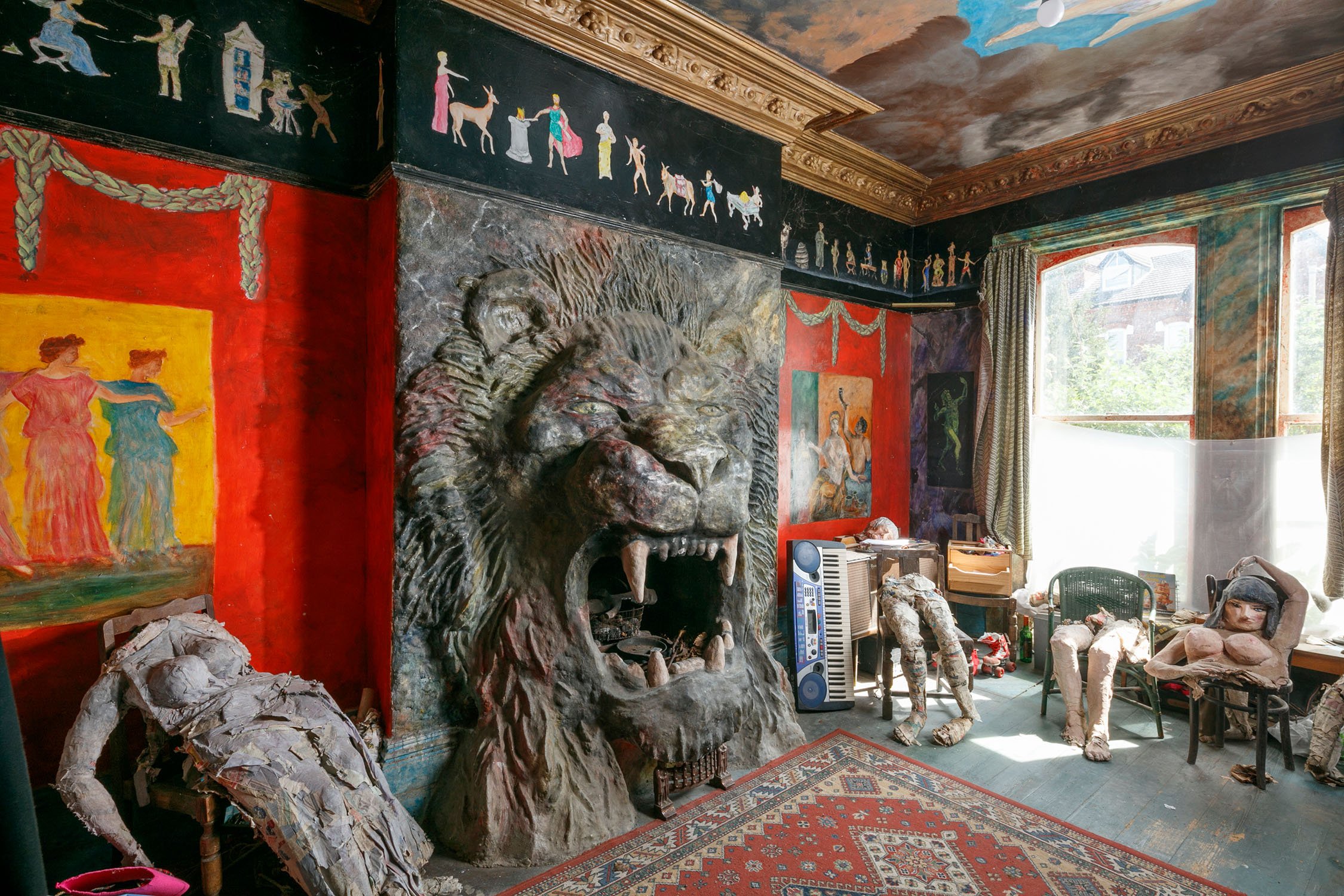 A photograph of a room containing a large lion head sculpture on the wall, a painted mural ceiling and partial models of people placed on chairs