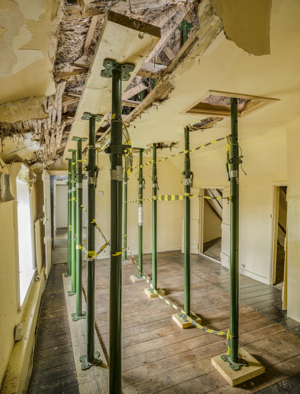 A series of metal poles standing on timber supports braced between the floor and a damaged ceiling.