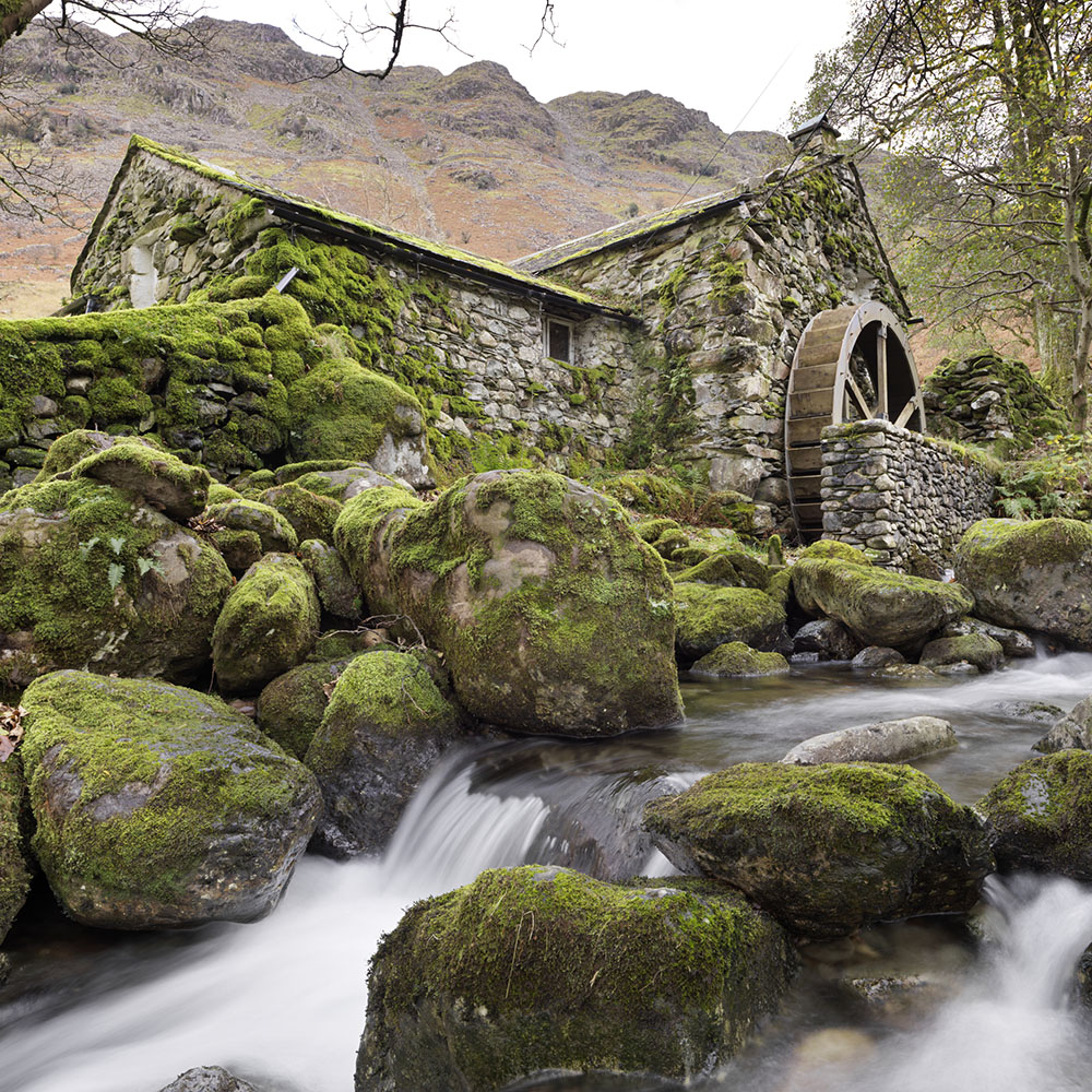 A stone-built water mill on a rocky stream in front of a tall hill.
