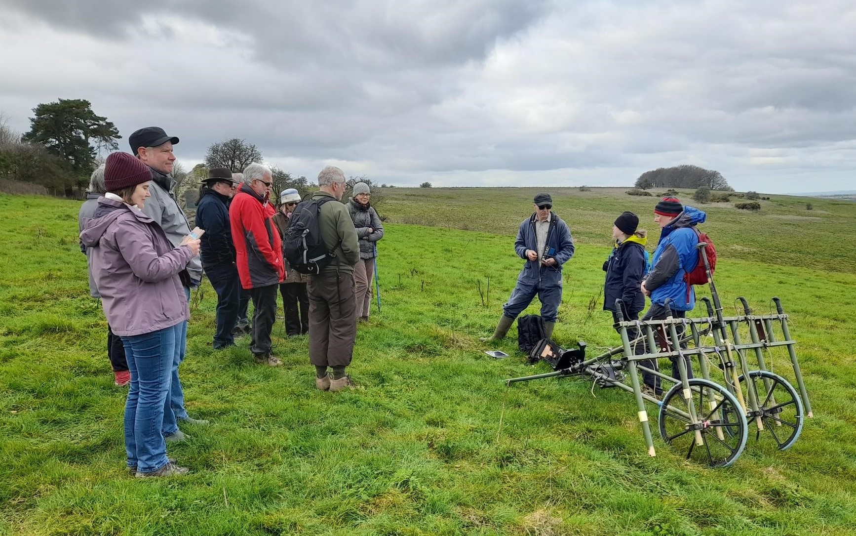 A photograph of a group of people in a field. They are listening to speaker who is demonstrating some archaeological survey equipment.
