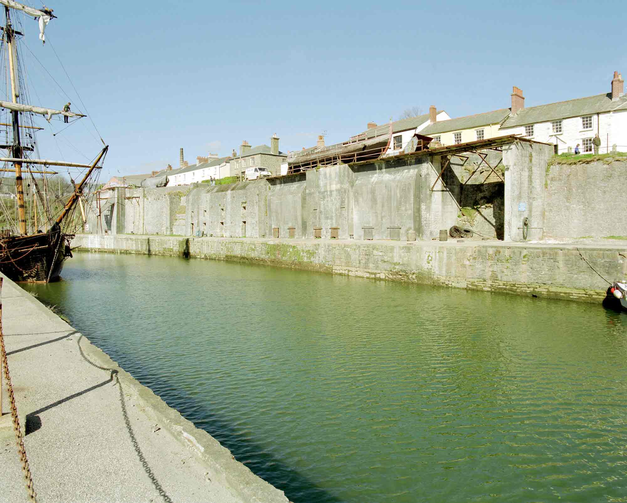 Sailing ship in harbour and houses can be seen beyond the high harbour walls