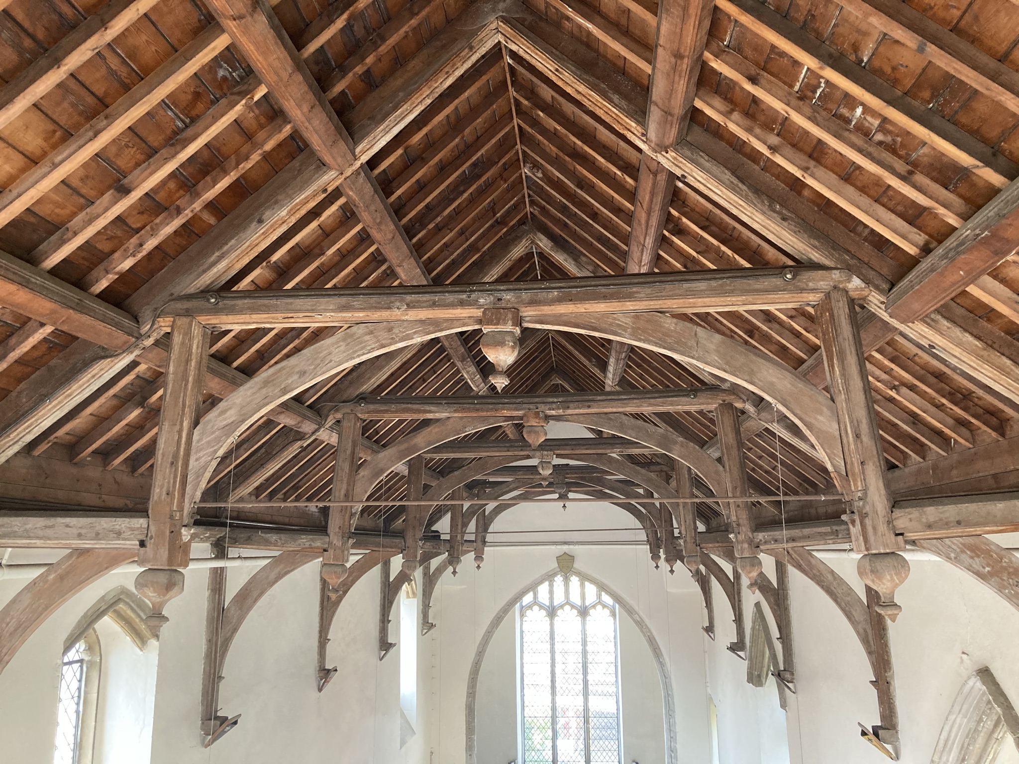 The wooden hammerbeam roof of Becket's Chapel can be seen. Wooden timbers with a series of central wooden arches. The arched plain glass chapel window can be seen in the background.