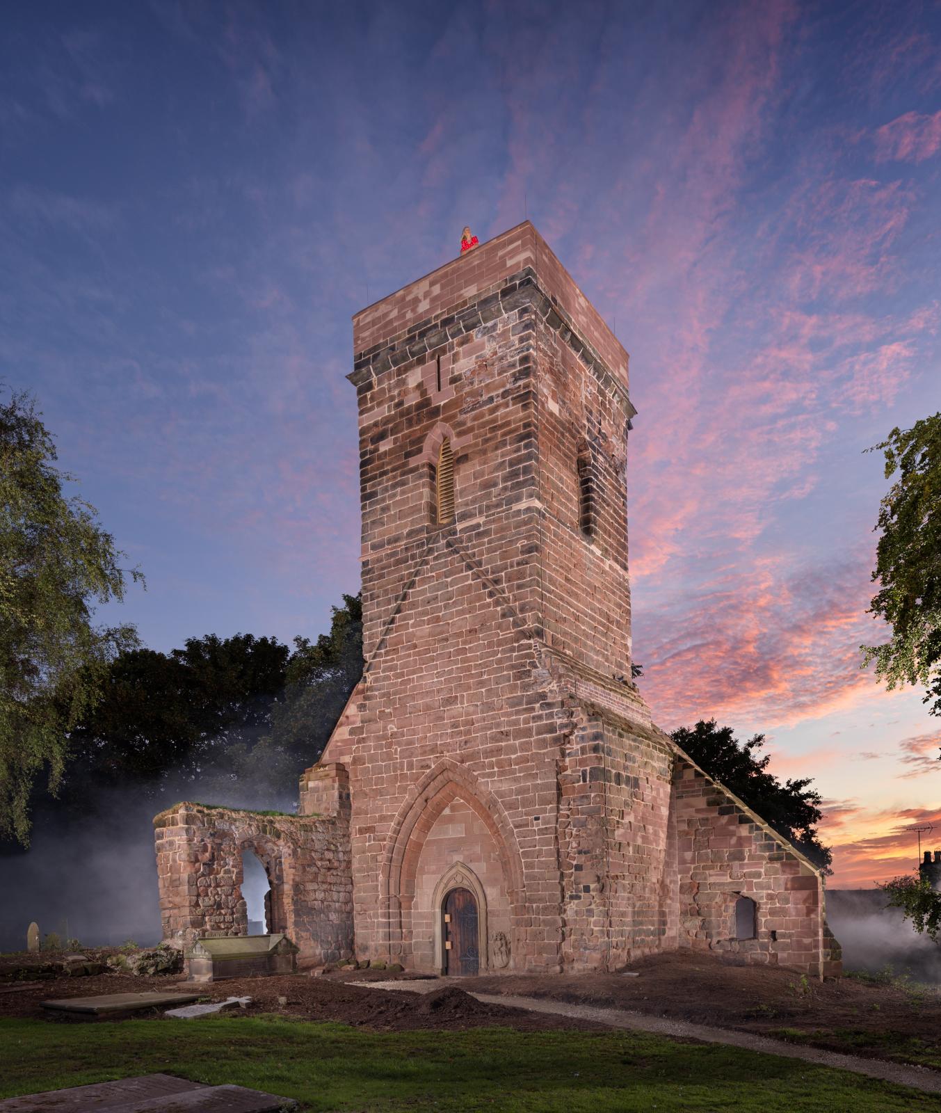The stone tower of a ruined church against a vivid sunset. A person in a red jacket stands at the top.