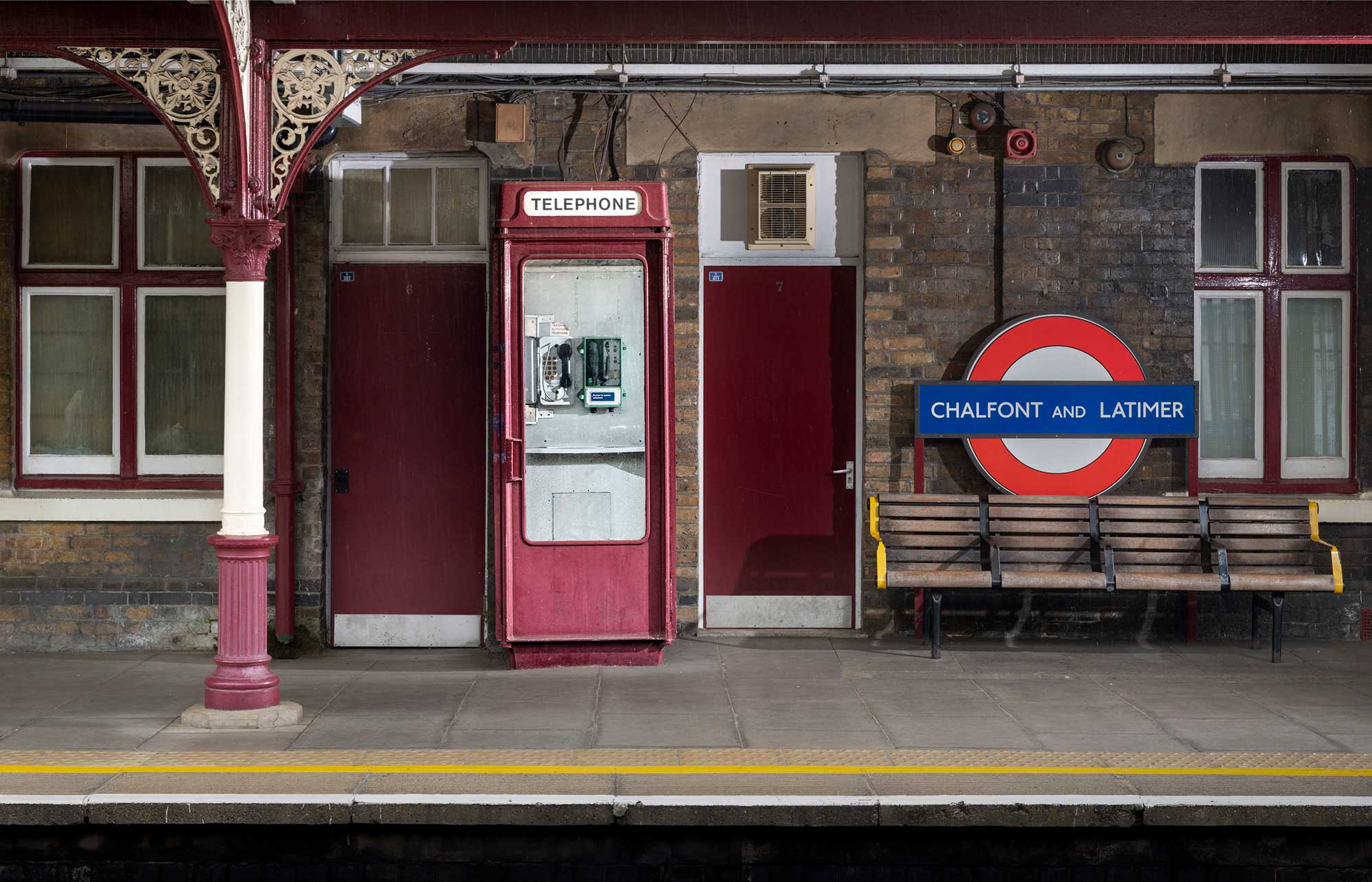 A telephone kiosk painted maroon on a railway station platform alongside seats.