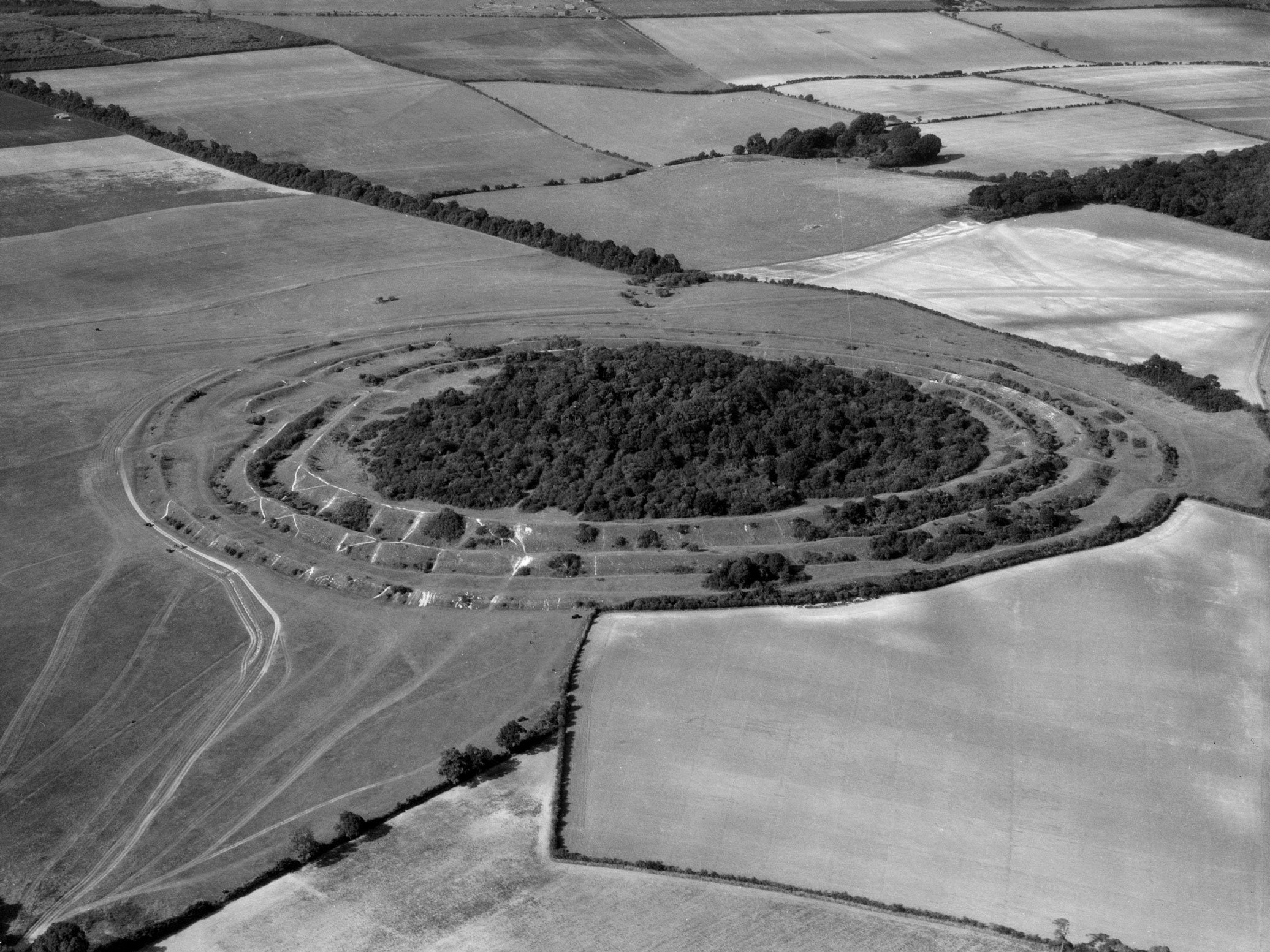 A black and white aerial photograph showing a hill fort with multiple banks and ditches: the interior is wooded.