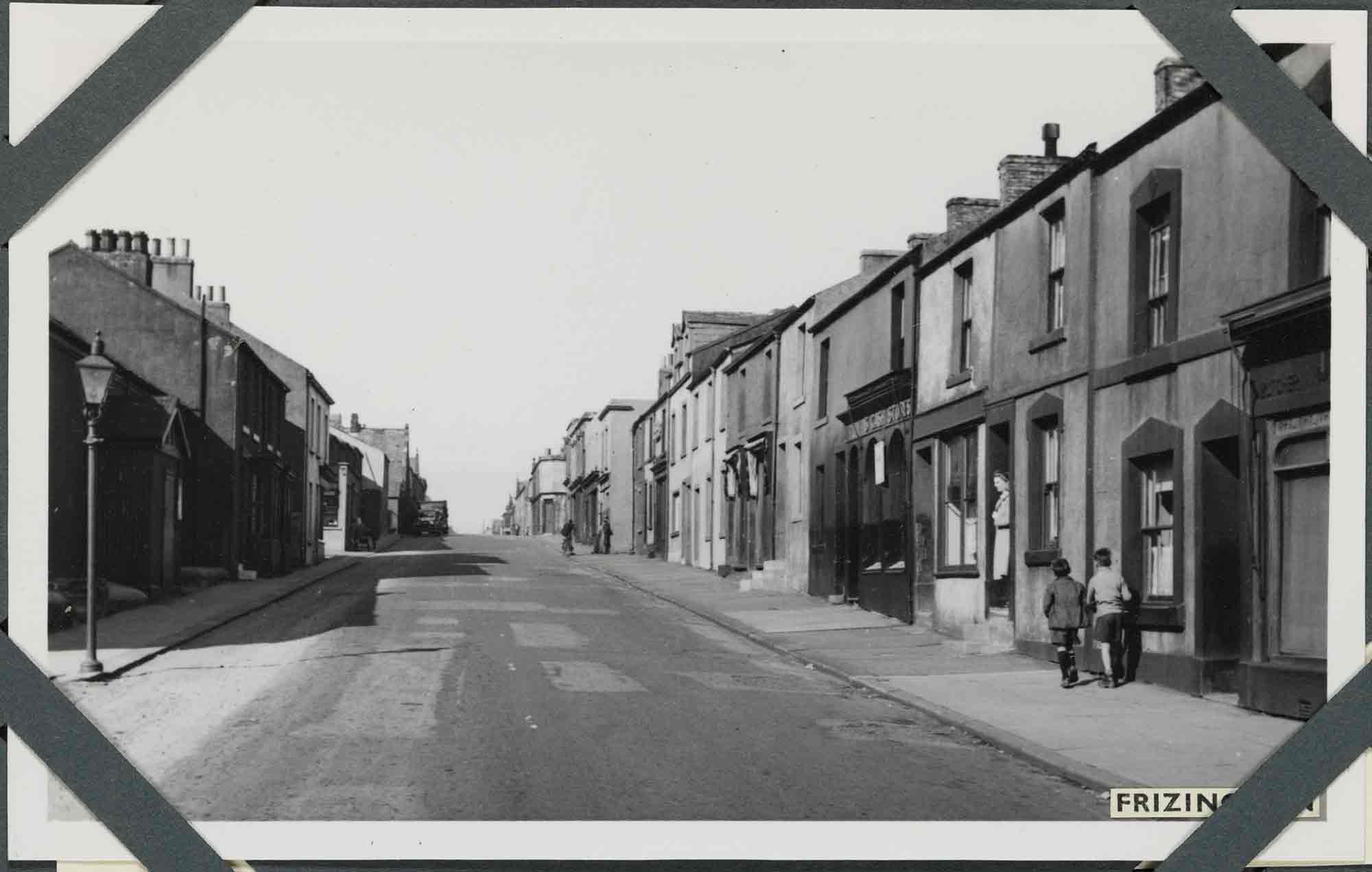Black and white photograph of an urban street scene. The view looks along a straight road that has a gentle incline. The road is flanked on either side by mainly two-storey houses and commercial premises that open directly onto the pavement. At the left of the photograph, a street lamp stands at the edge of the pavement. On the opposite side, two boys walk side-by-side away from the camera, approaching a woman standing on a doorstep. A label applied to the bottom-right of the photograph reads: 'Frizington', part of which is obscured by one of four corner photograph album mounts.