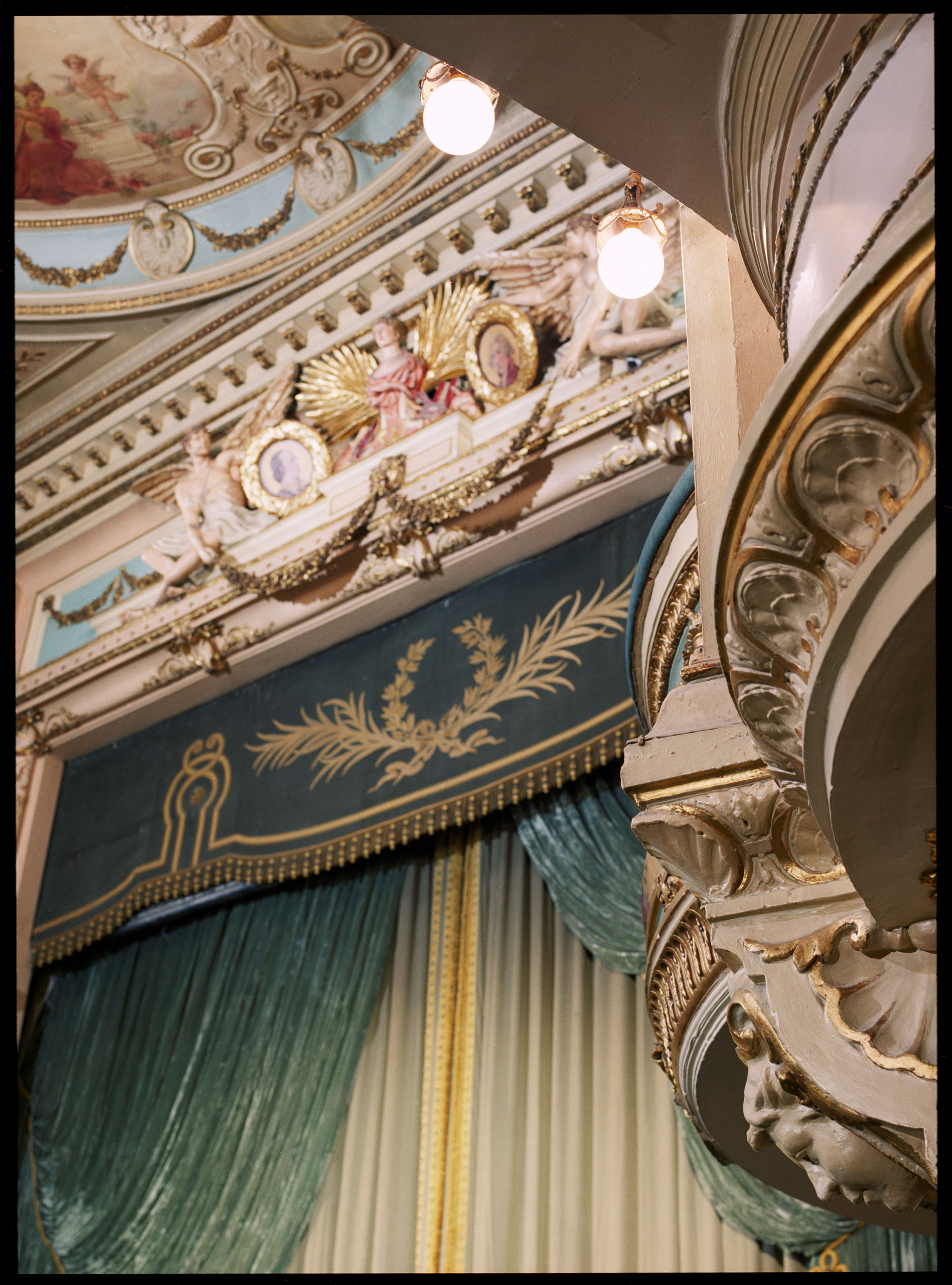 Interior of a theatre showing colourful plaster decoration to the stage area and box fronts with classically inspired motifs.
