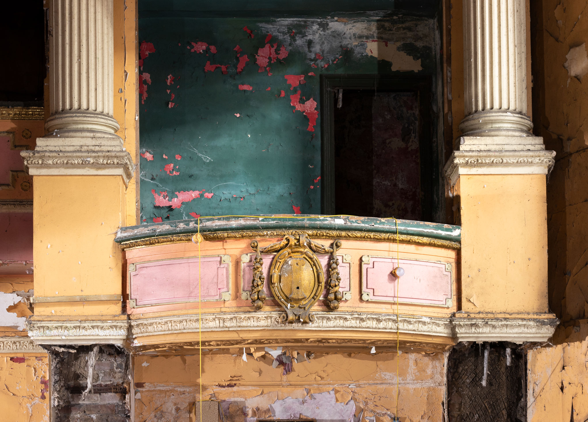 Detail of an ornate but derelict theatre interior, a balcony front with decorative moulding, flanked by columns.
