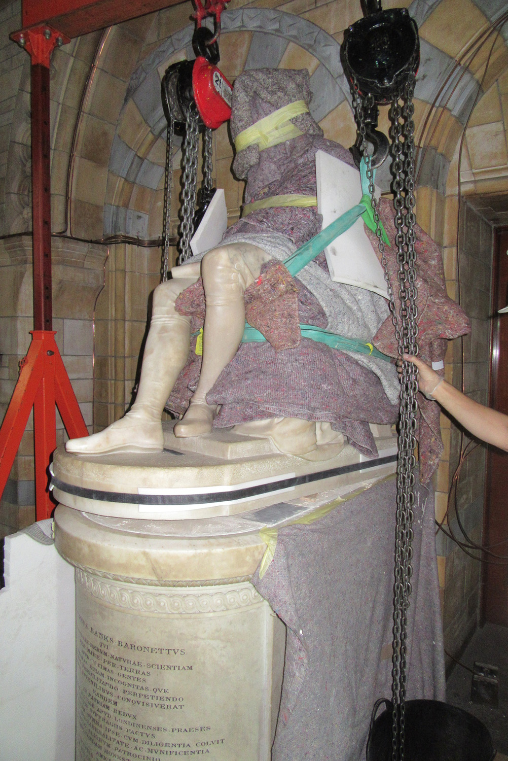 A stone statue of a seated figure covered in blankets and foam padding is lifted by two winches.