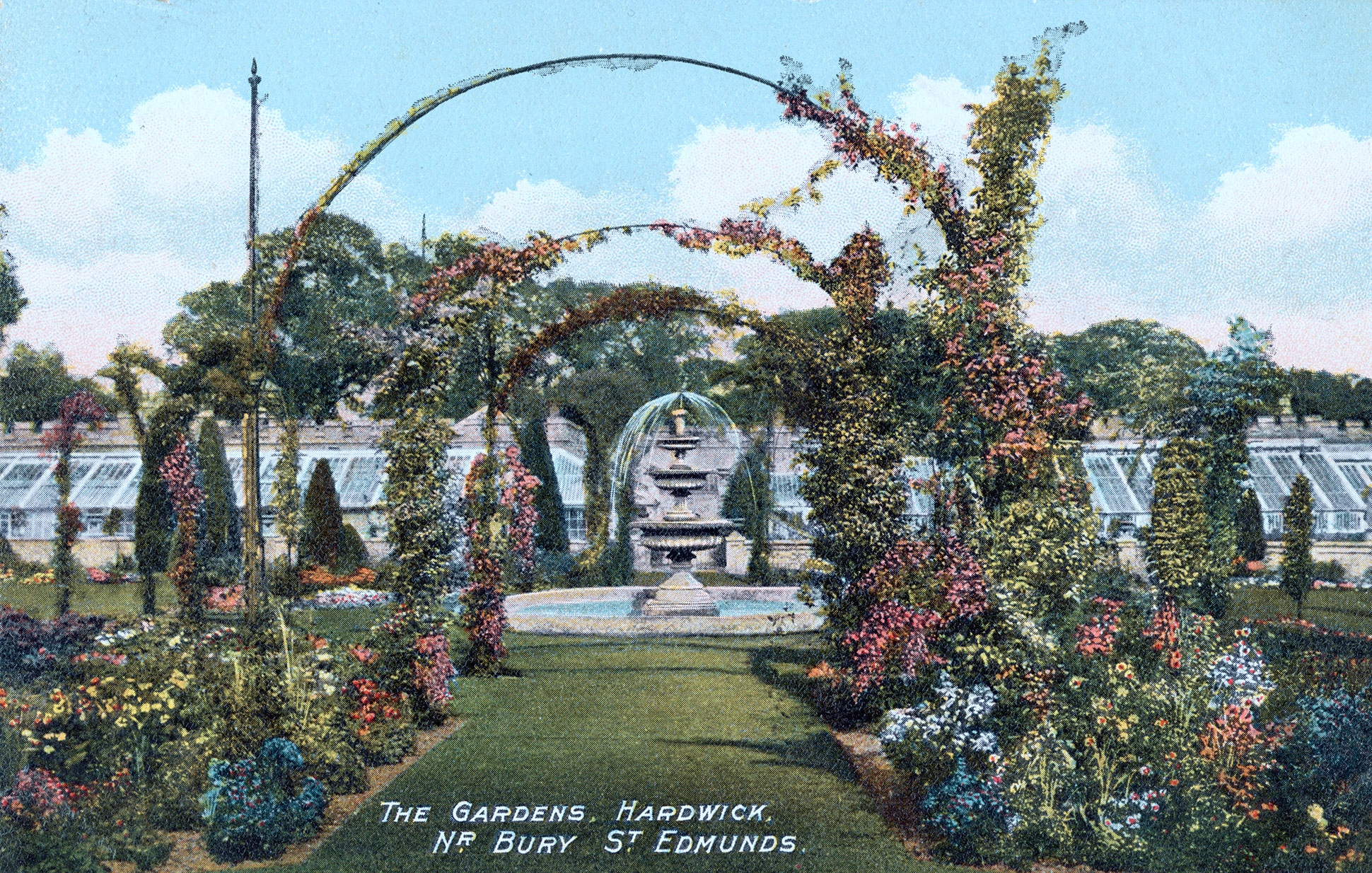 A colourised historic postcard shows a central fountain with arches in the foreground and flower borders either side of a grass path. Glass houses can be seen in the background.