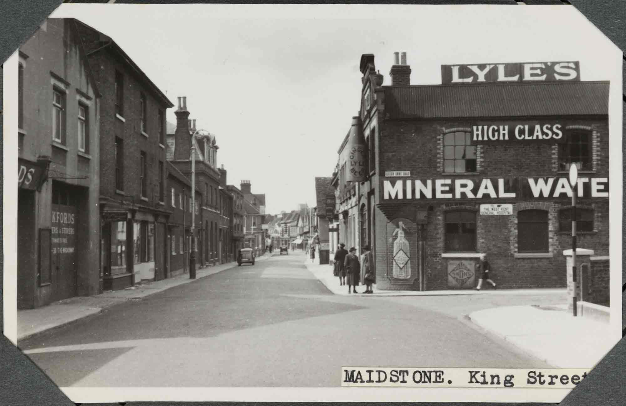 Black and white photograph of an urban street scene featuring two- and three-storey houses and commercial premises. A few people stand or walk along the pavement to the right of the near-empty street. Two women in long coats and hats stand on the corner of the street, outside a brick building that carries signs for 'Lyle's High Class Mineral Wate' [Water]. Above them, suspended from the first floor of the building is an oversized bottle. A label applied to the bottom-right of the photograph reads: 'Maidstone. King Street', part of which is obscured by one of four corner photograph album mounts. 