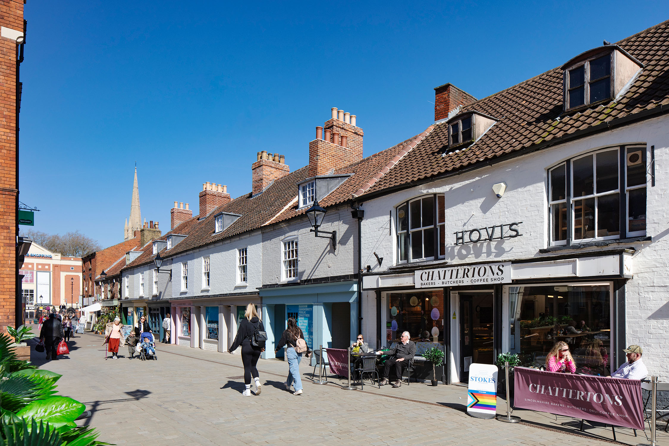 An urban street scene of shops and people on a high street in Lincoln, UK. Signs on the coffee shop in the foreground read 'Hovis / Chattertons'.