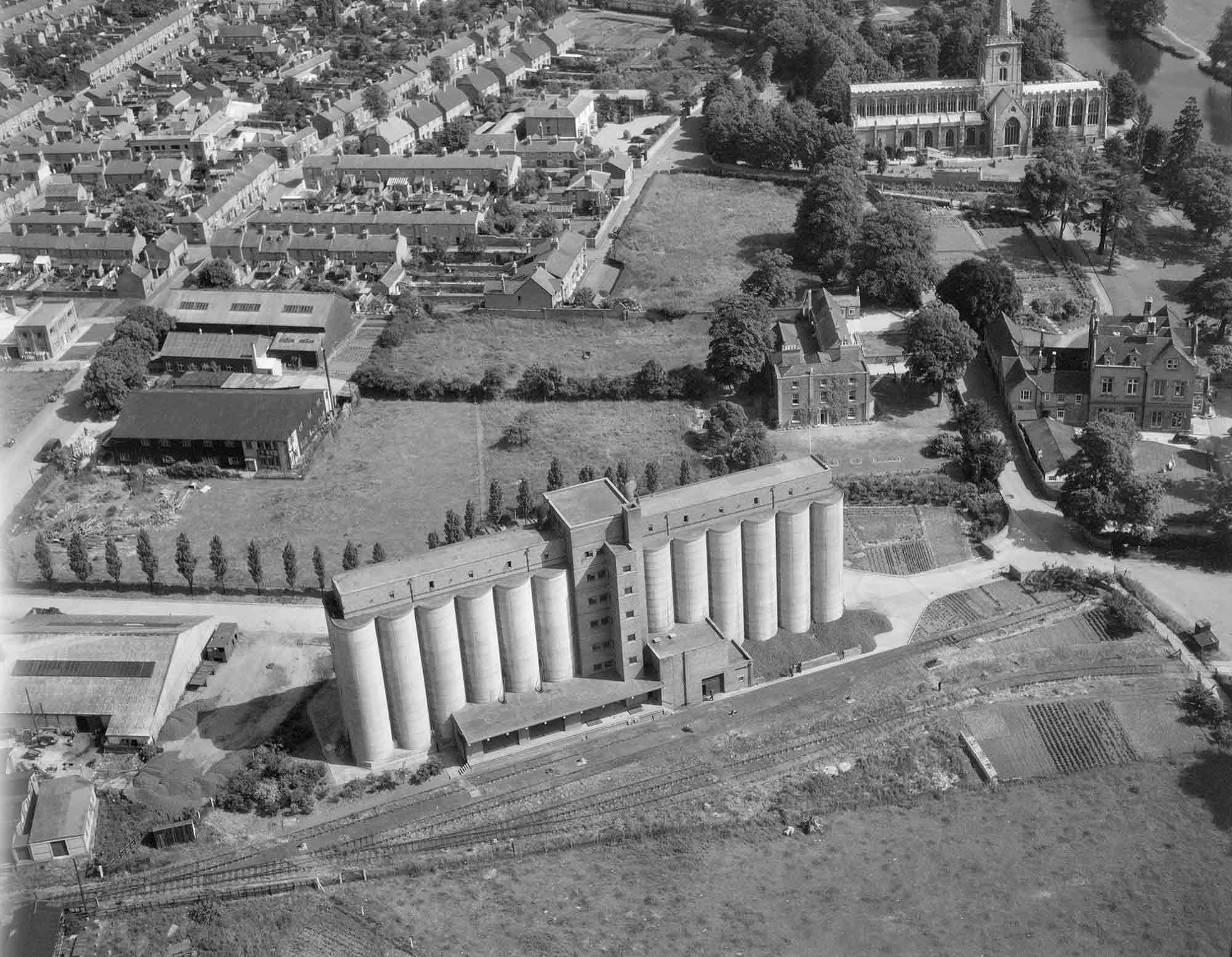 Black and white oblique aerial photograph of a concrete grain silo. The tall silo, which has twelve upright cylinder-like segments to its front, stands in the middle foreground. It is fronted by a railway siding and another track that crosses the front of the photograph. Behind and to the right of the silo are two large, detached houses, and behind to the left are streets with various domestic and other buildings. In the top-right corner is a large gothic church with a tower topped with a spire that is truncated by the edge of the photograph.