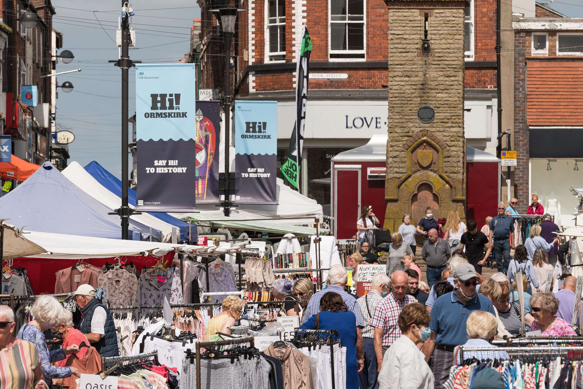 A busy high street market with clothes handing on rails under marquees. Banners hang from the lamp posts which say Hi! Ormskirk – Say Hi! to History