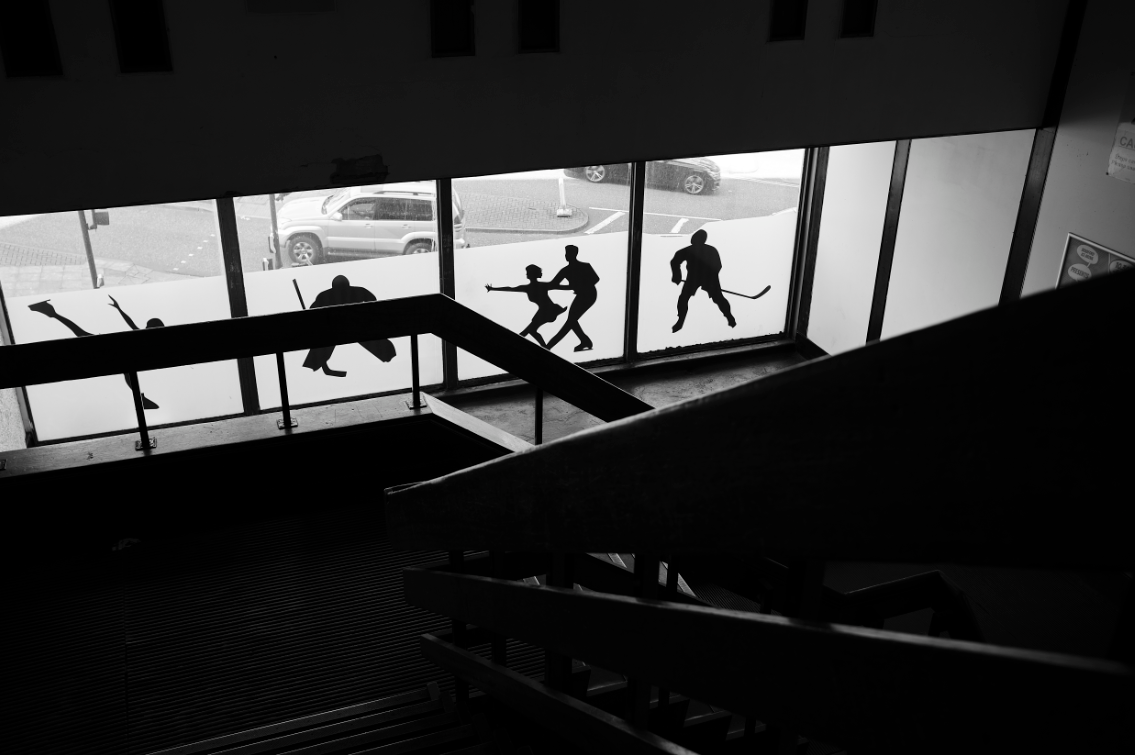 Black and white photograph taken from the staircase of a building. The windows are decorated with different images of skaters, including an ice hockey player and two figure skaters.