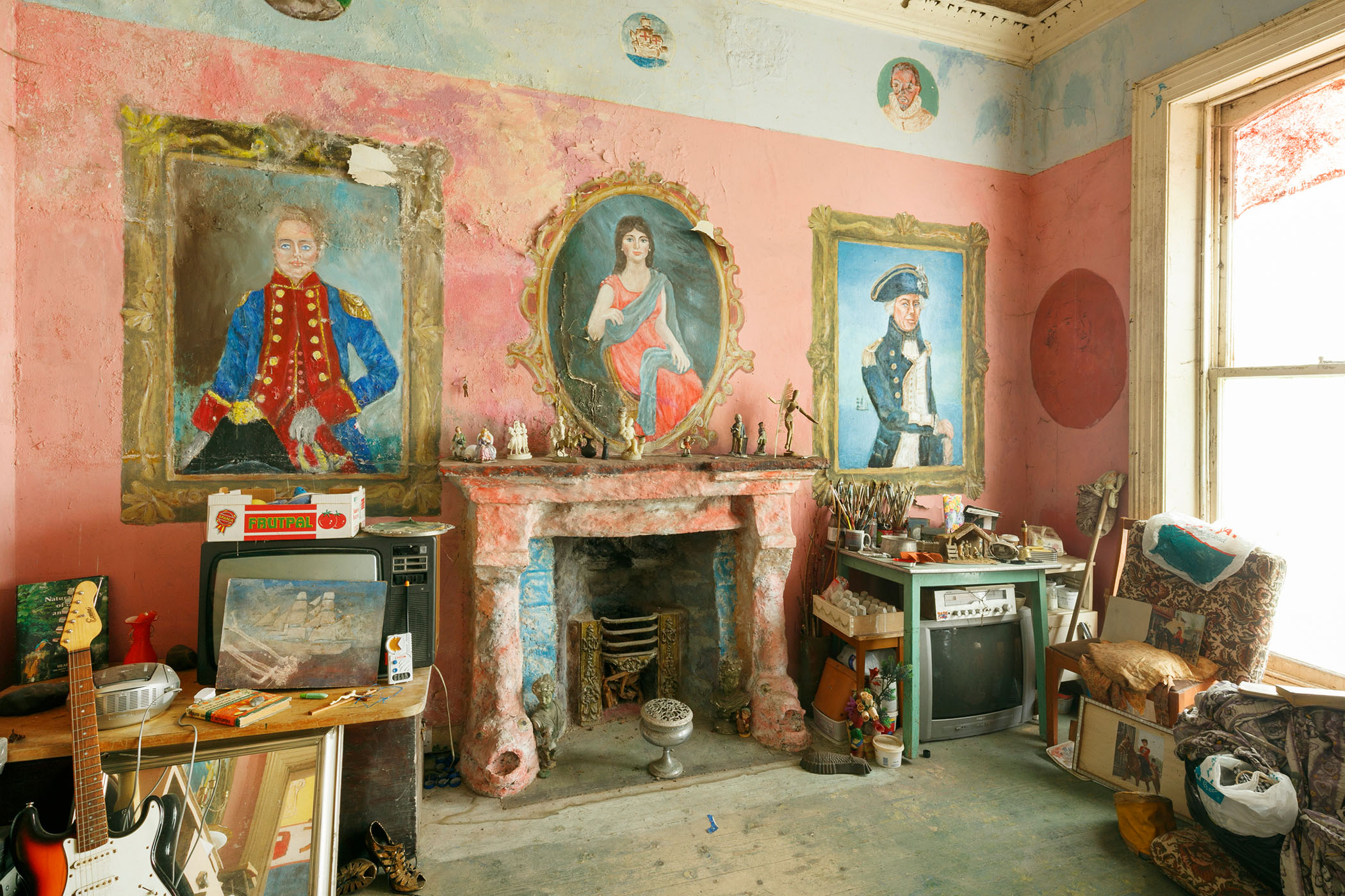 A photograph of a room containing an old, disused fireplace and 3 figures within decorative frames painted on the wall