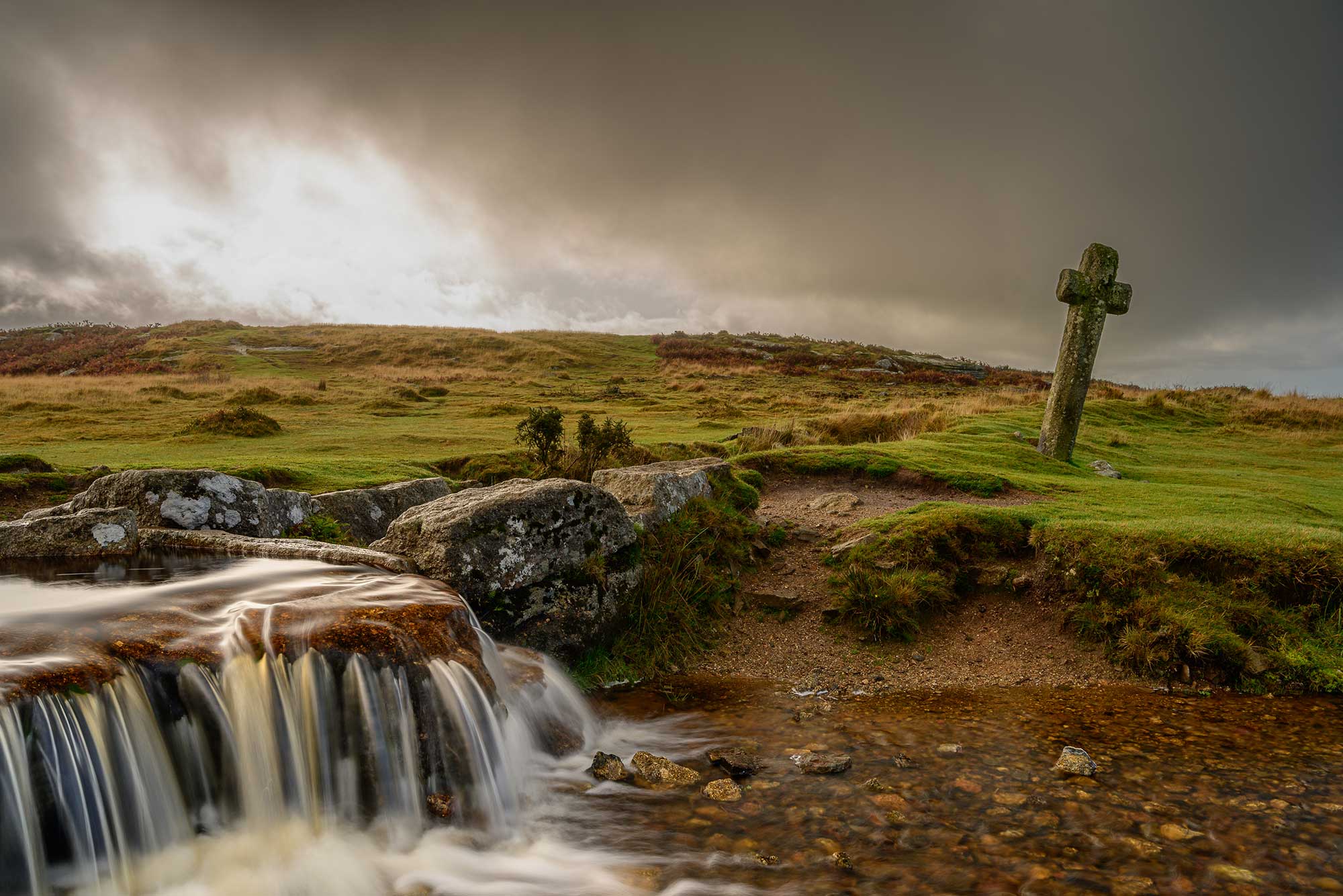A photograph of a moorland landscape with a small waterfall flowing into a stream, and a vertical stone cross positioned alongside the water.