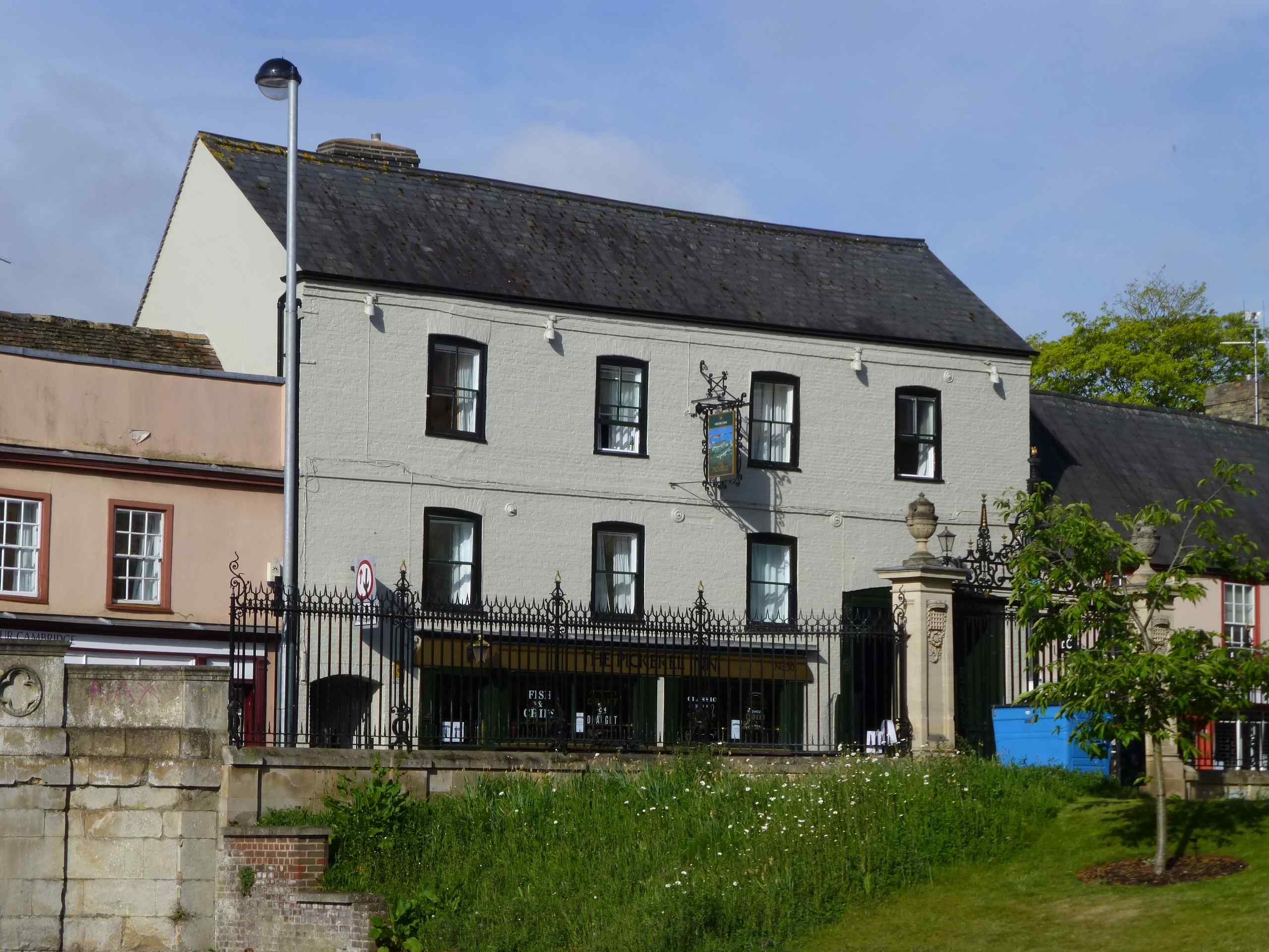 Wide shot of the exterior of the Pickerel Inn in Cambridge, UK. A stone wall, black iron fence and grass bank are in the lower half of the image between the building and the photographer.