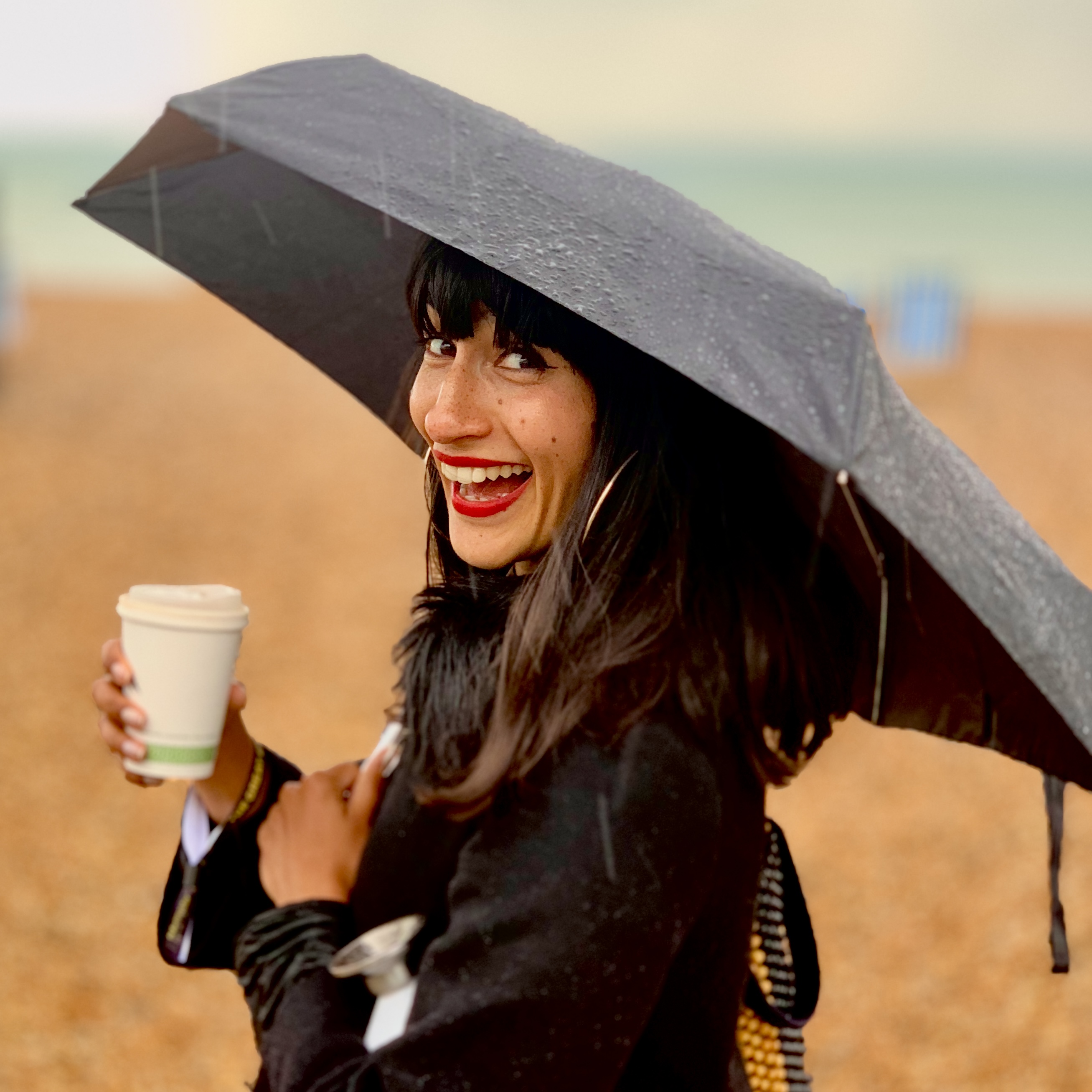 A portrait photograph of Susie Thornberry on a beach smiling while holding an umbrella and a takeaway coffee cup