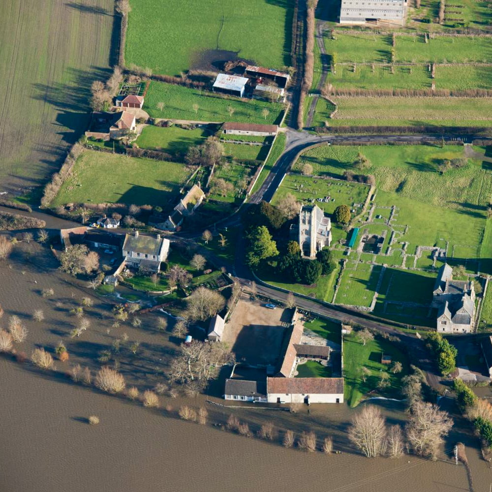 Birds-eye view of a village with bare trees and building foundations. Greyish-brown water has flooded the bottom half of the image. 