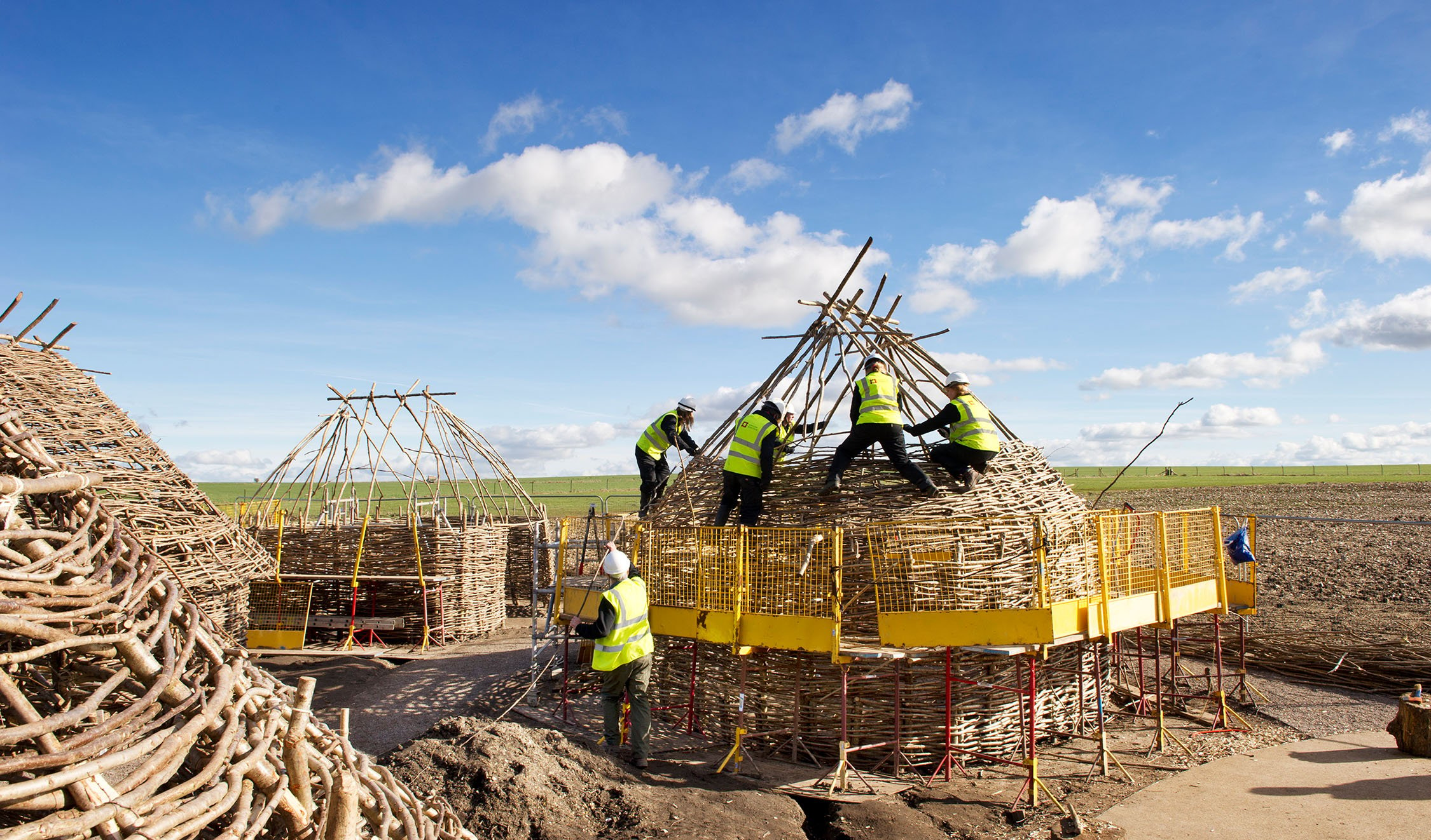 Five people in high-visibility clothing and helmets constructing a Neolithic-style hut.
