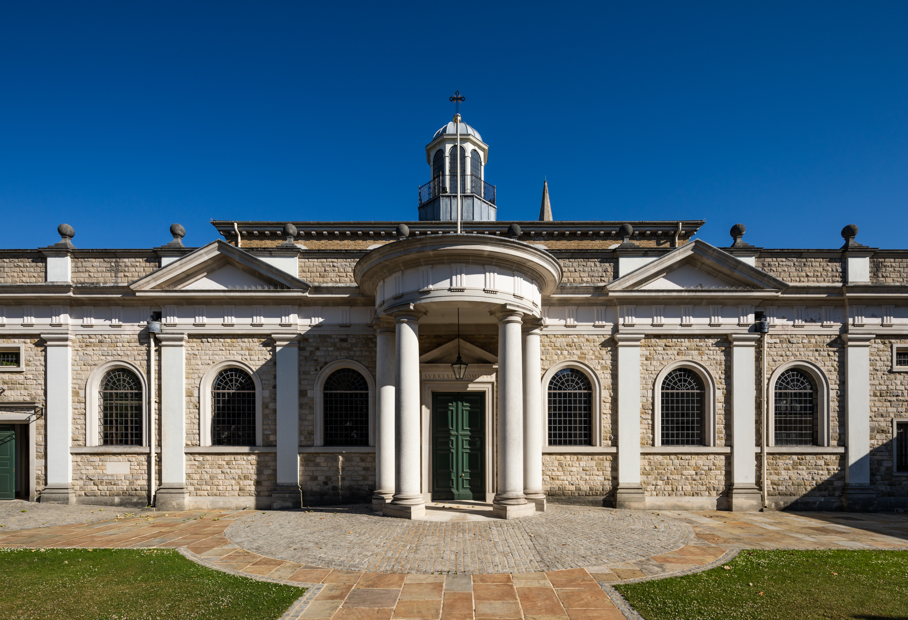 The exterior of Brentwood Cathedral shows a cobbled and paved circular external flooring. A rounded porch with pillars and a long line exterior wall with semi-circular leaded windows and two arches, with a decorative hexagonal tower.