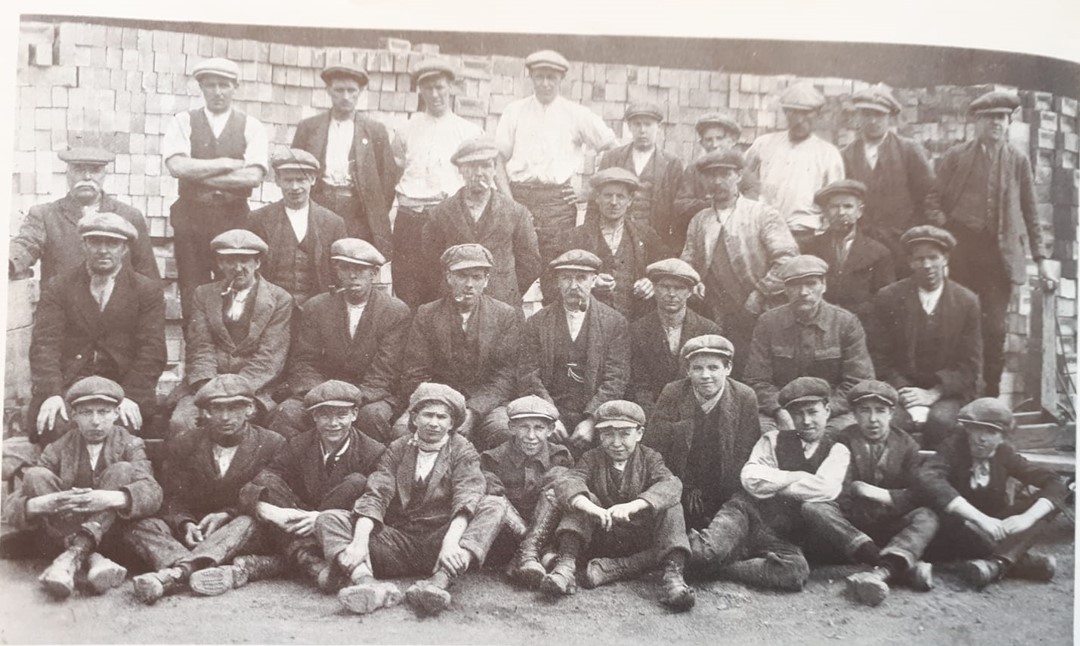 A black and white photo of rows of men and boys in working clothes, wearing flat caps.