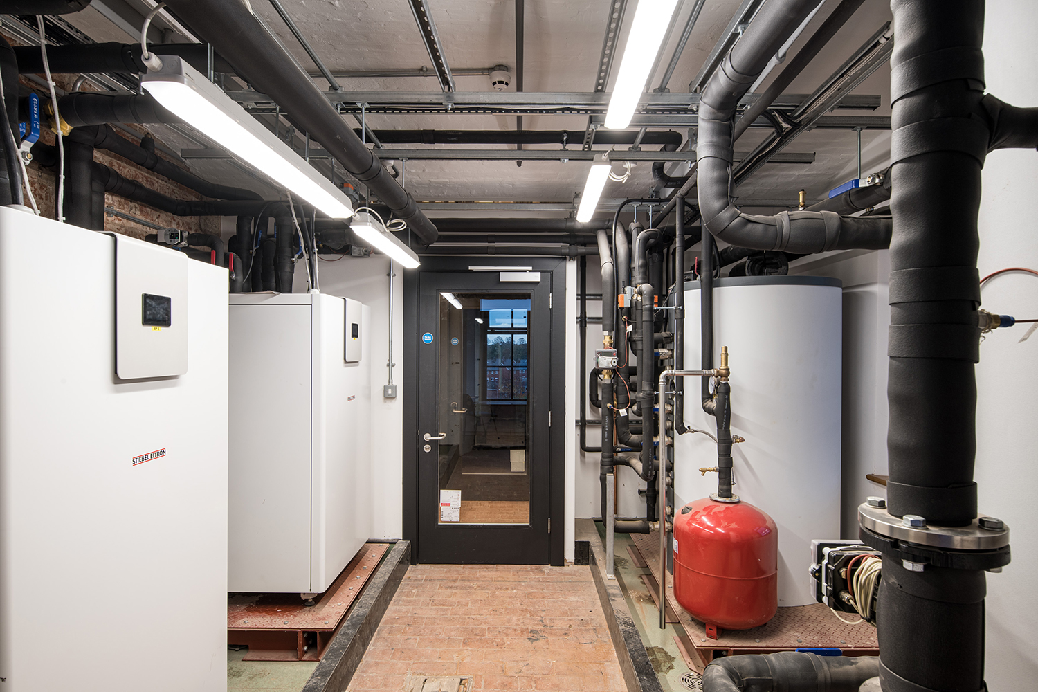 A central walk way with white boxed units on the left, a tall white cylinder and small red cylinder on the right, and black  pipes.
