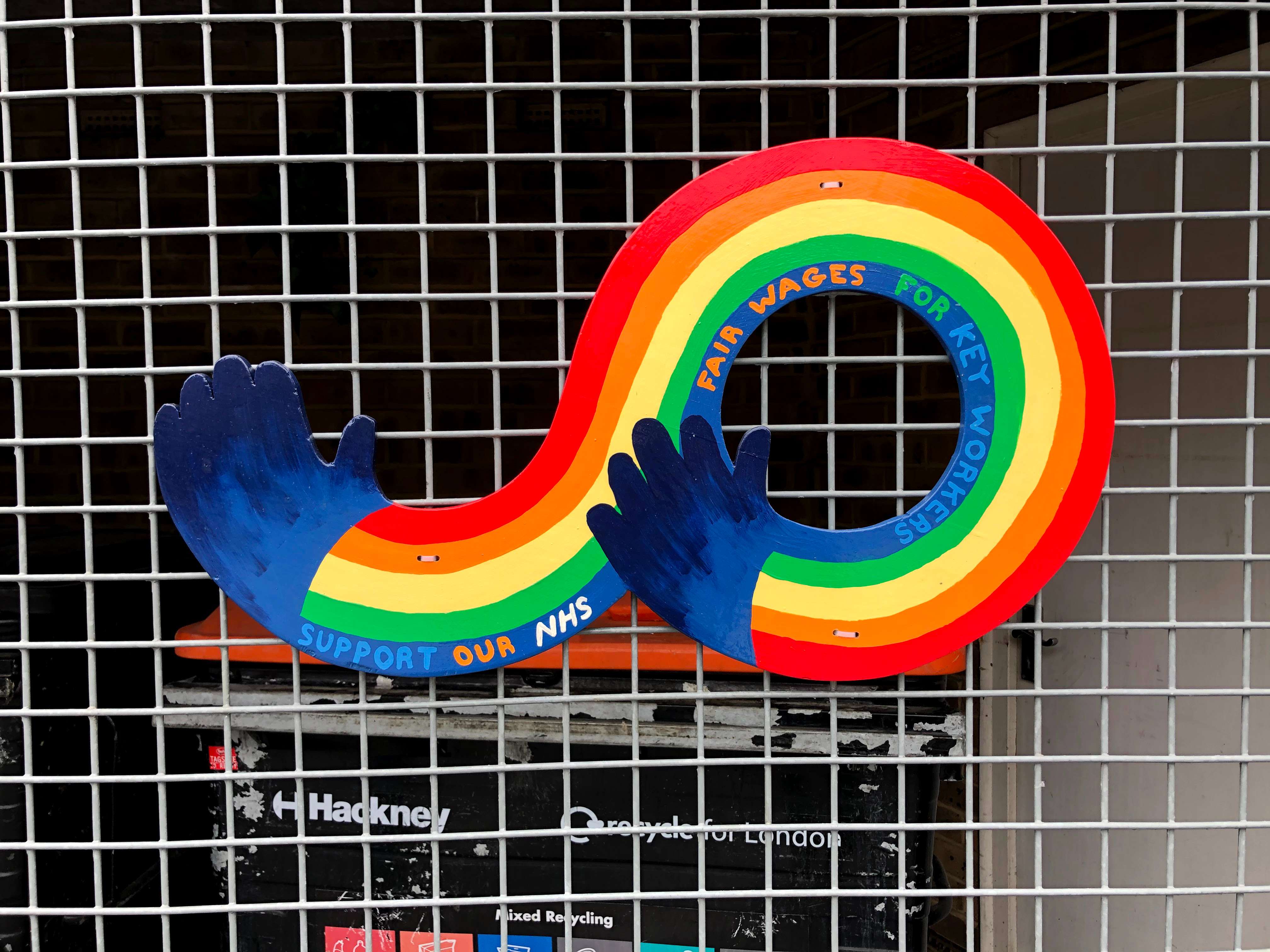 Rainbow colours with hands at each. Words 'Support Our NHS, Fair Wages for Key Workers' written on the blue band of the rainbow. The graphic is attached to a metal fence.