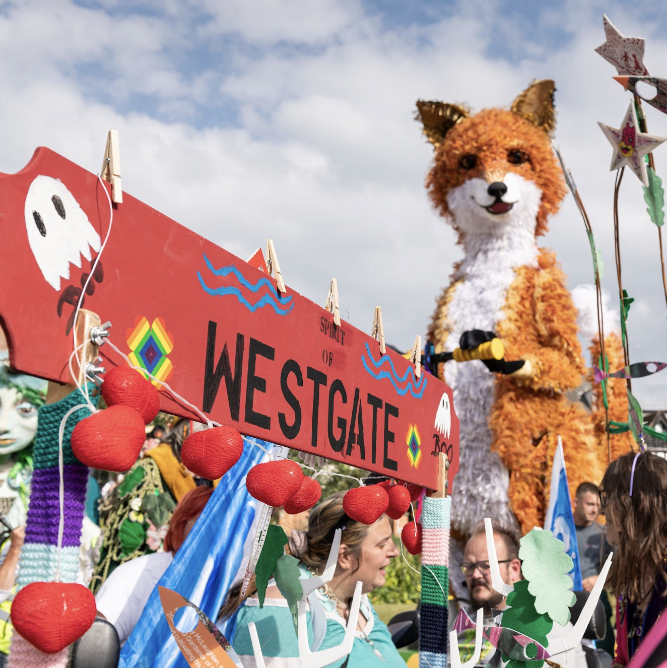 A carnival street scene with crowds, a close-up of a sign reading 'Spirit of Westgate' in the foreground, and a large fox puppet in the background.