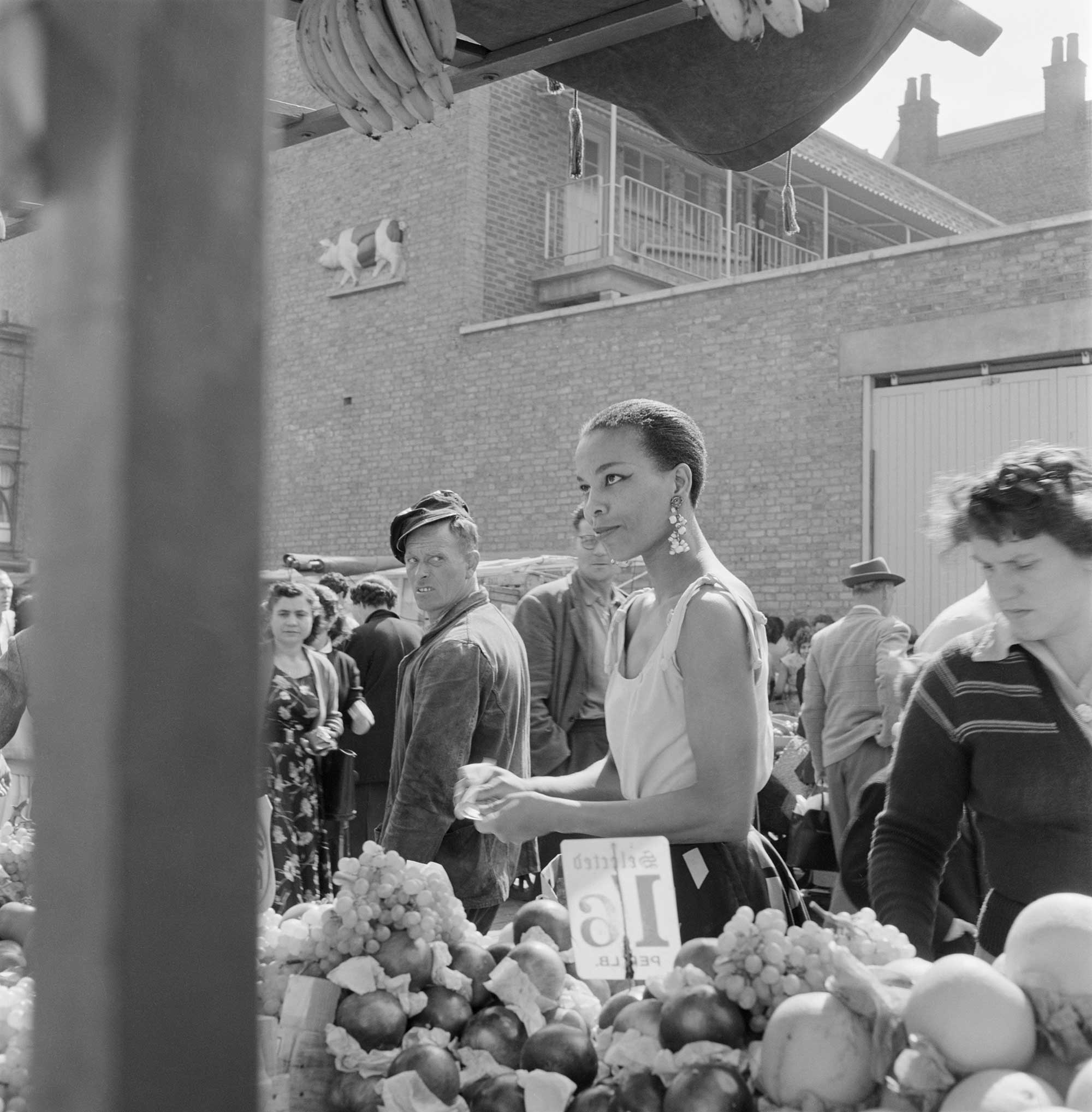 An informal portrait of a young black woman buying fruit at an unidentified open air market in north london, with a man looking back over his shoulder at her.