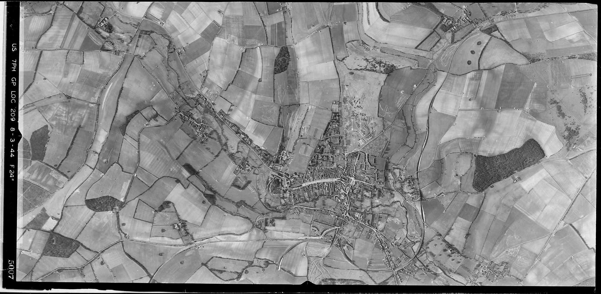 A black and white vertical aerial photograph of an urban area set in a landscape of fields and valleys. The settlement includes a wide central street with plots stretching back from it. Patches of woodland are dotted amongst the patchwork of fields.