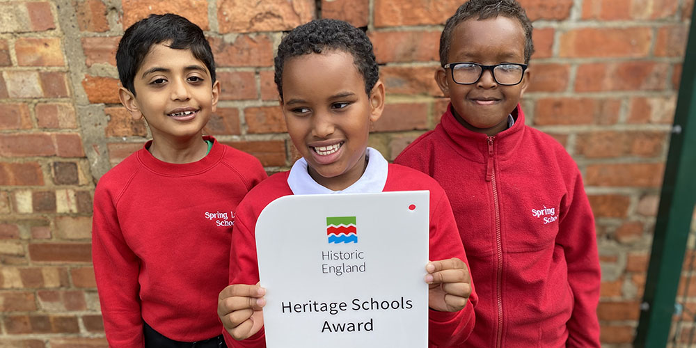 Three children in red school jumpers stand in front of a brick wall, holding up a plaque that reads "Historic England: Heritage Schools Award".