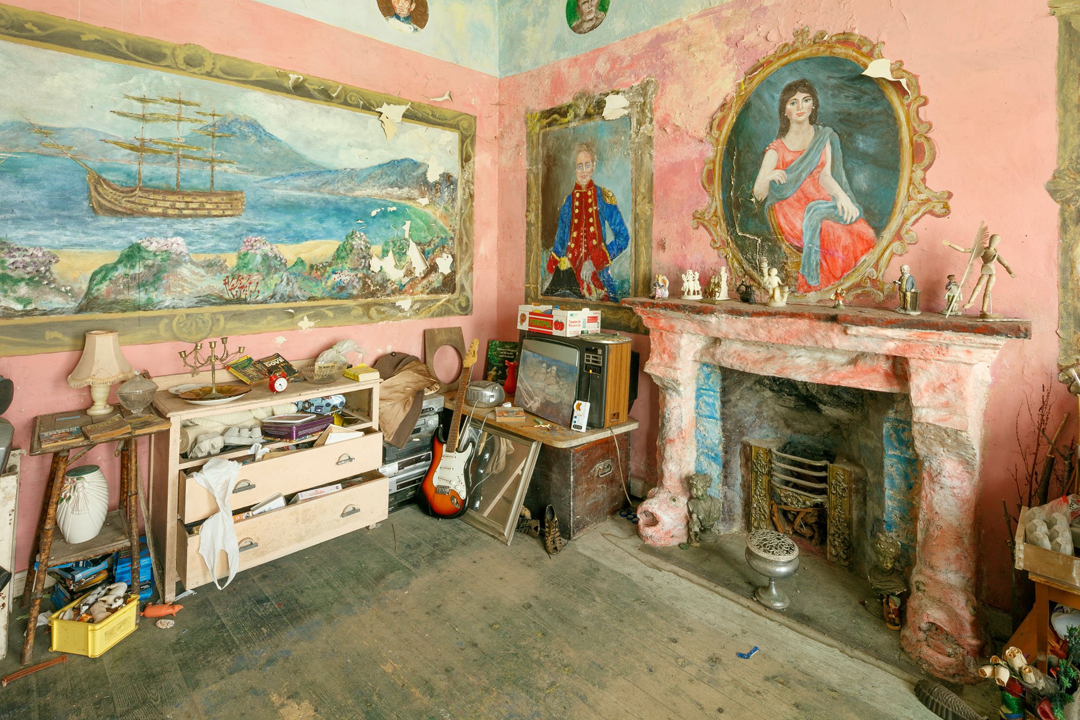 A photograph of a room containing an old fireplace, and wall paintings of two figures within decorative frames and a ship within a coastal setting.
