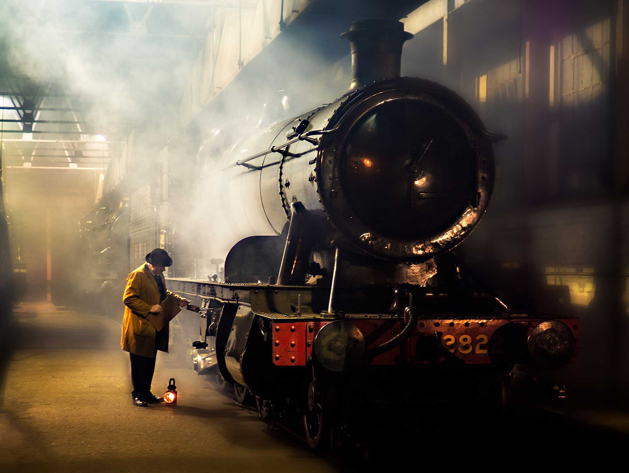 A photograph of a steam engine in a railway station, with a person conducting mechanical inspections of the engine.