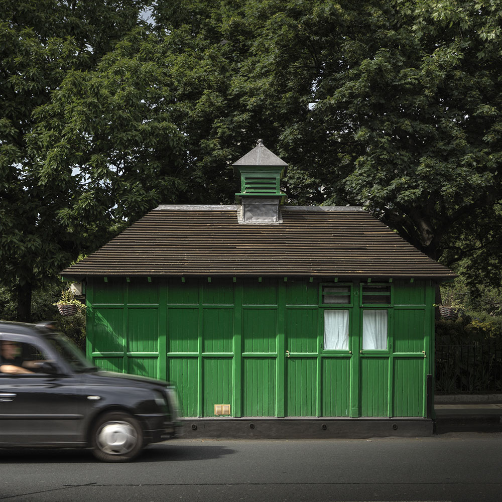 A small green wooden building with a small cote on the roadside with a modern black cab driving by.