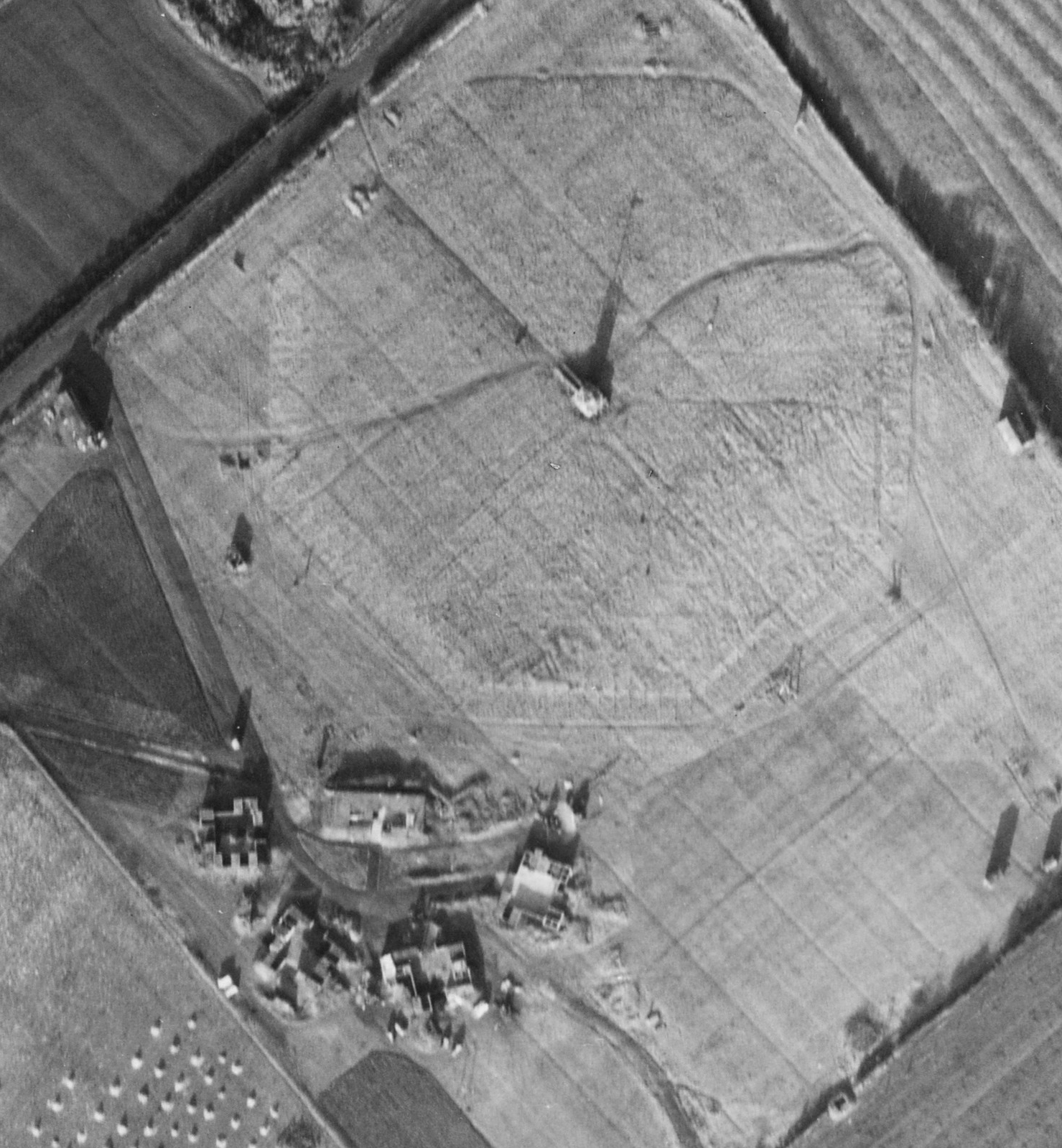 A black and white vertical aerial photograph of an octagonal shape set within a field. In the centre of the octagon is a structure that gives a tall, slender shadow. To the bottom-left are some buildings.