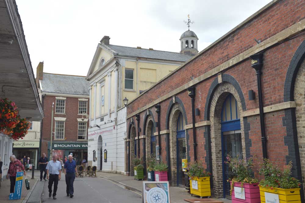 A photograph looking up a historic high street. A long brick arcaded building on the right is a covered market. It joins onto the Guildhall, a white, classical building in the middle ground.